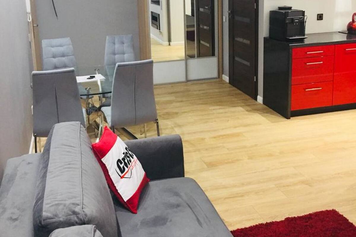 Una sala de estar moderna con un sofá gris, un cojín rojo y blanco y una alfombra roja. Hay una zona de comedor con una mesa de vidrio y sillas, y una cocina con armarios rojos y una encimera negra.