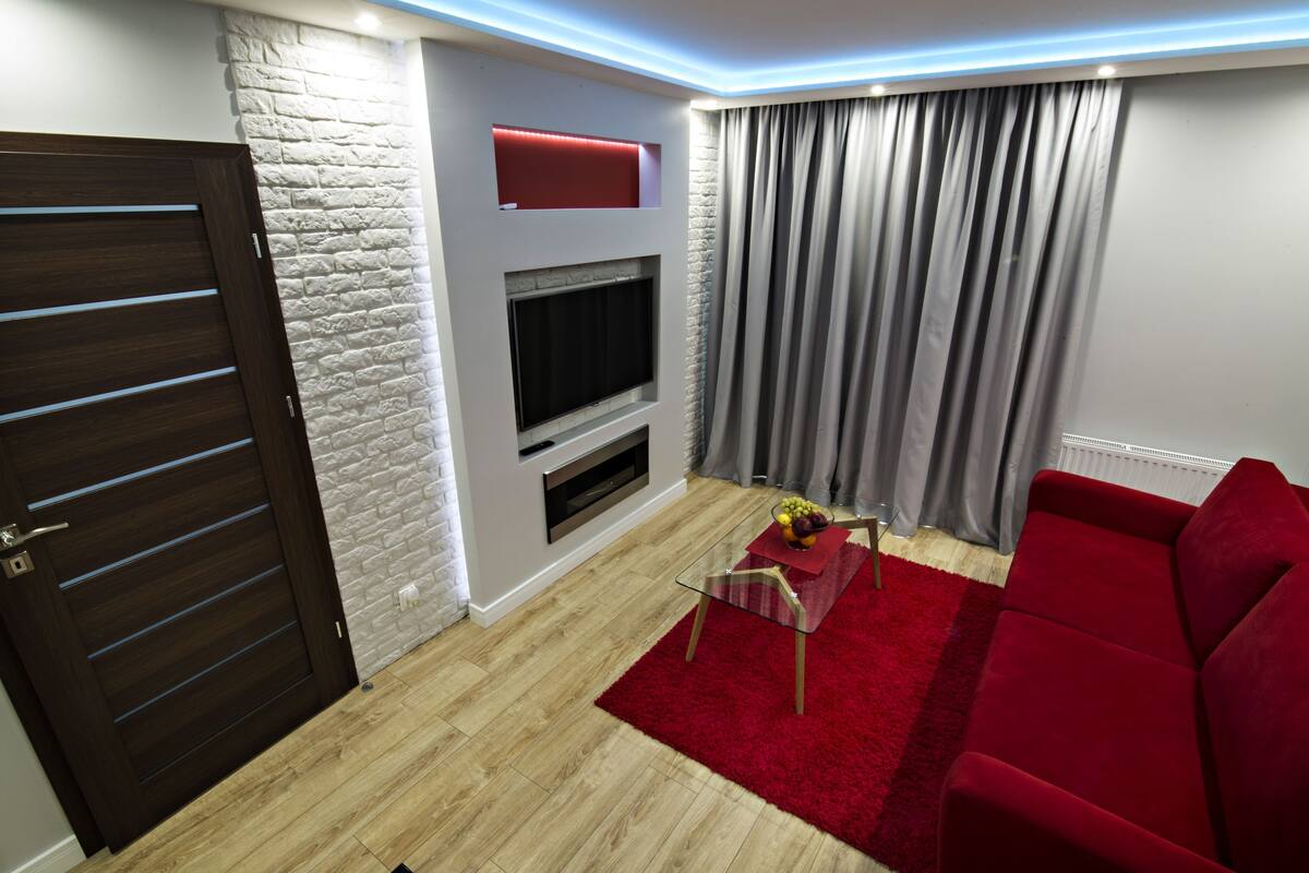 Una sala de estar con un sofá rojo, una mesa de centro de vidrio, un televisor montado en una pared de ladrillo blanco y una puerta de madera.