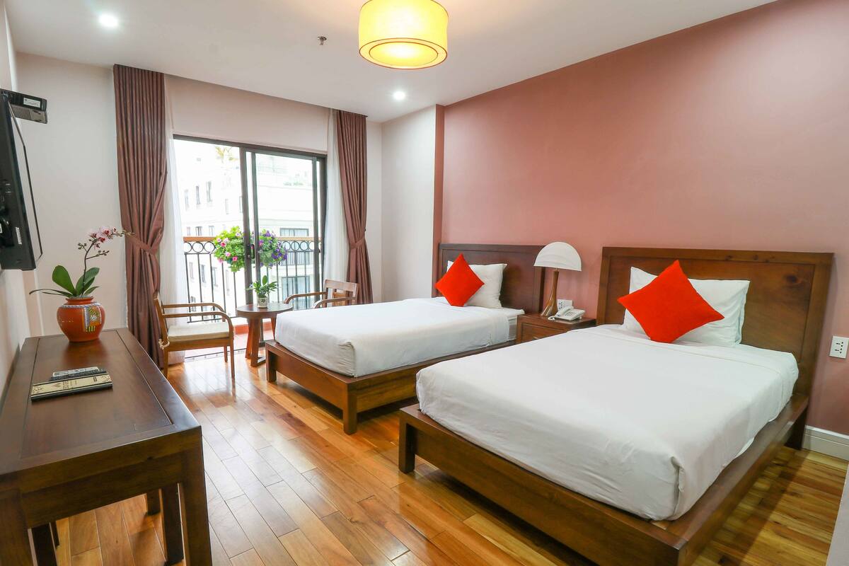 Een hotelkamer met twee bedden, een bureau, een stoel, een bijzettafeltje met een plant en uitzicht op buiten.