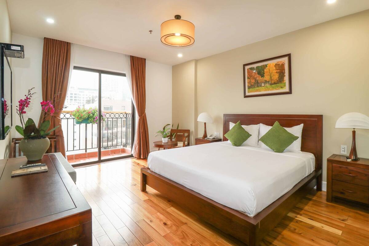 Een hotelkamer met een houten bed, een bureau, een stoel, een klein tafeltje met een plant en een balkon met gordijnen.