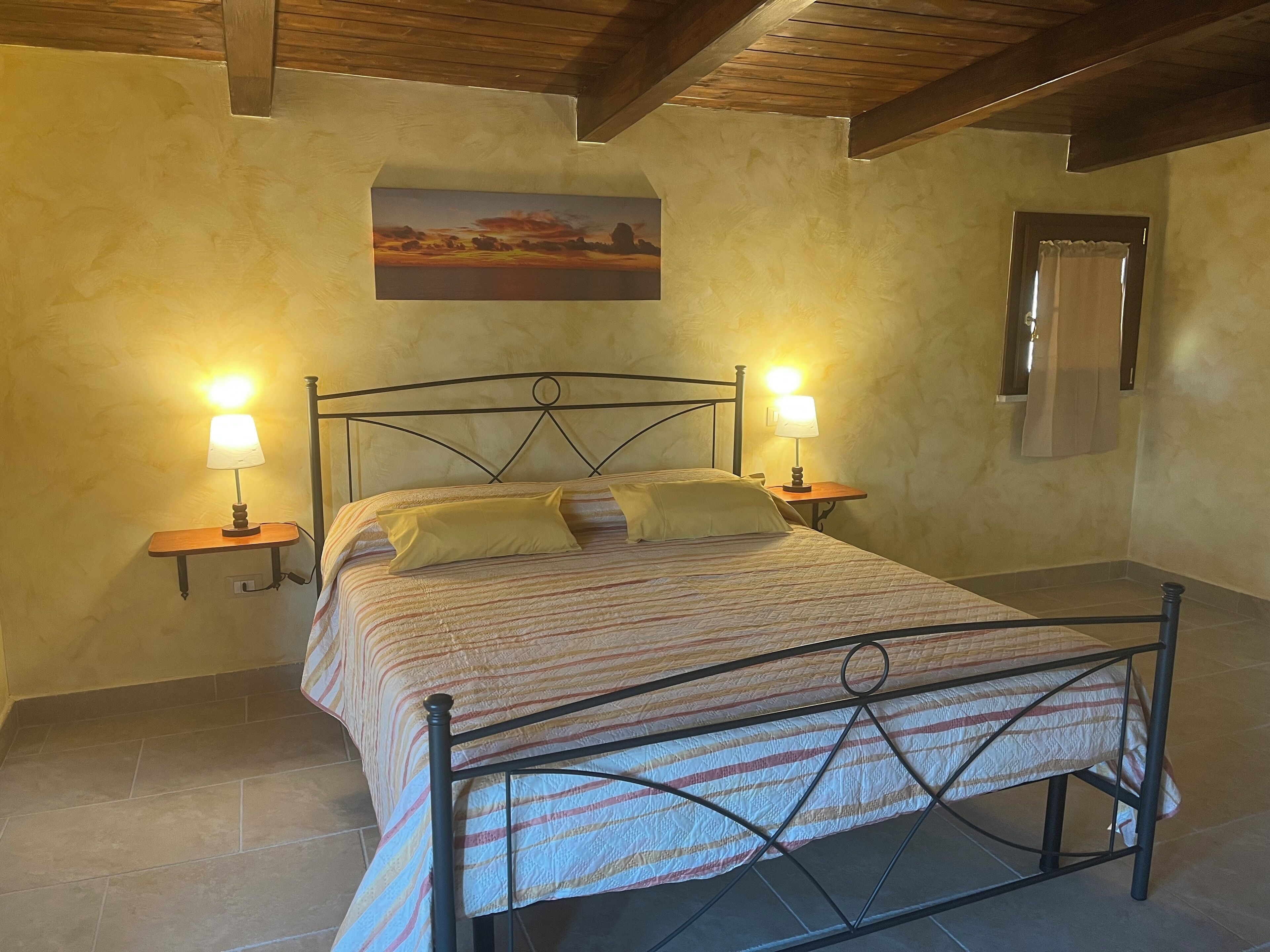 A bedroom with a bed, two bedside lamps, a painting on the wall, and a window with curtains.