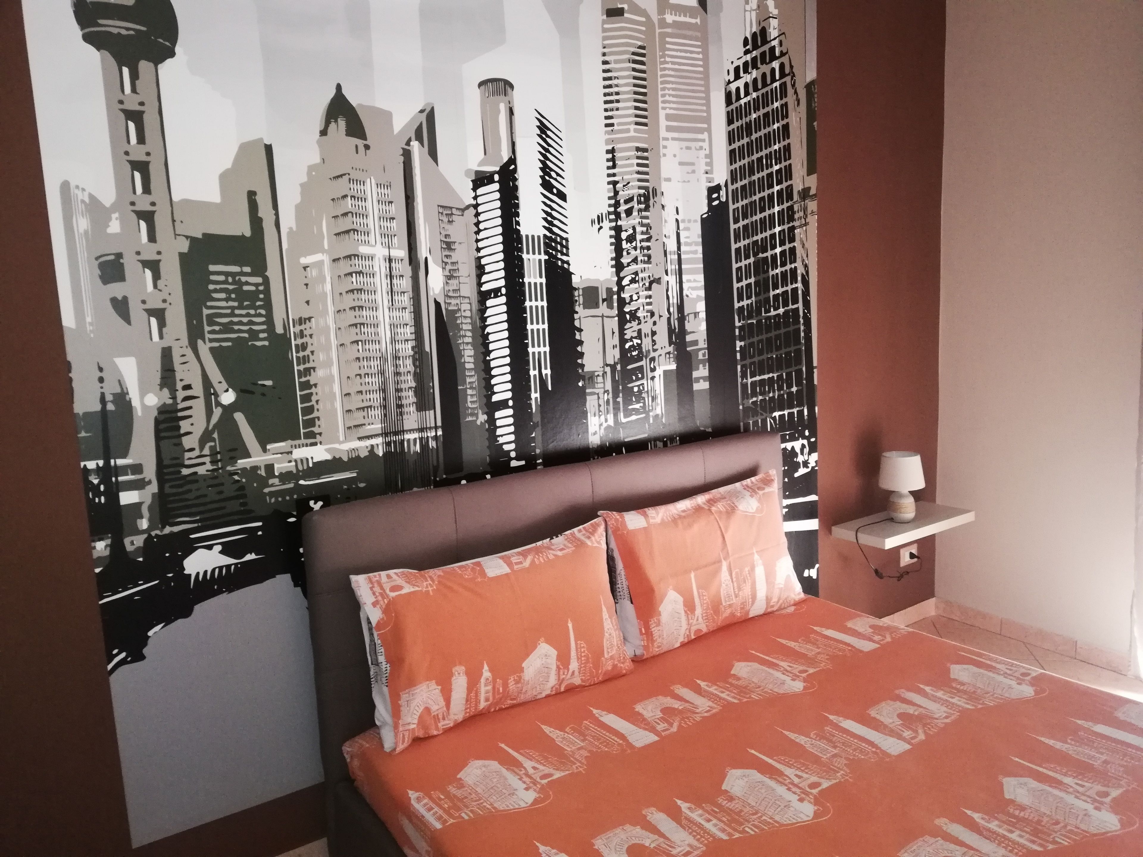 A bedroom with a cityscape mural on the wall, a bed with orange bedding, and a bedside table with a lamp.