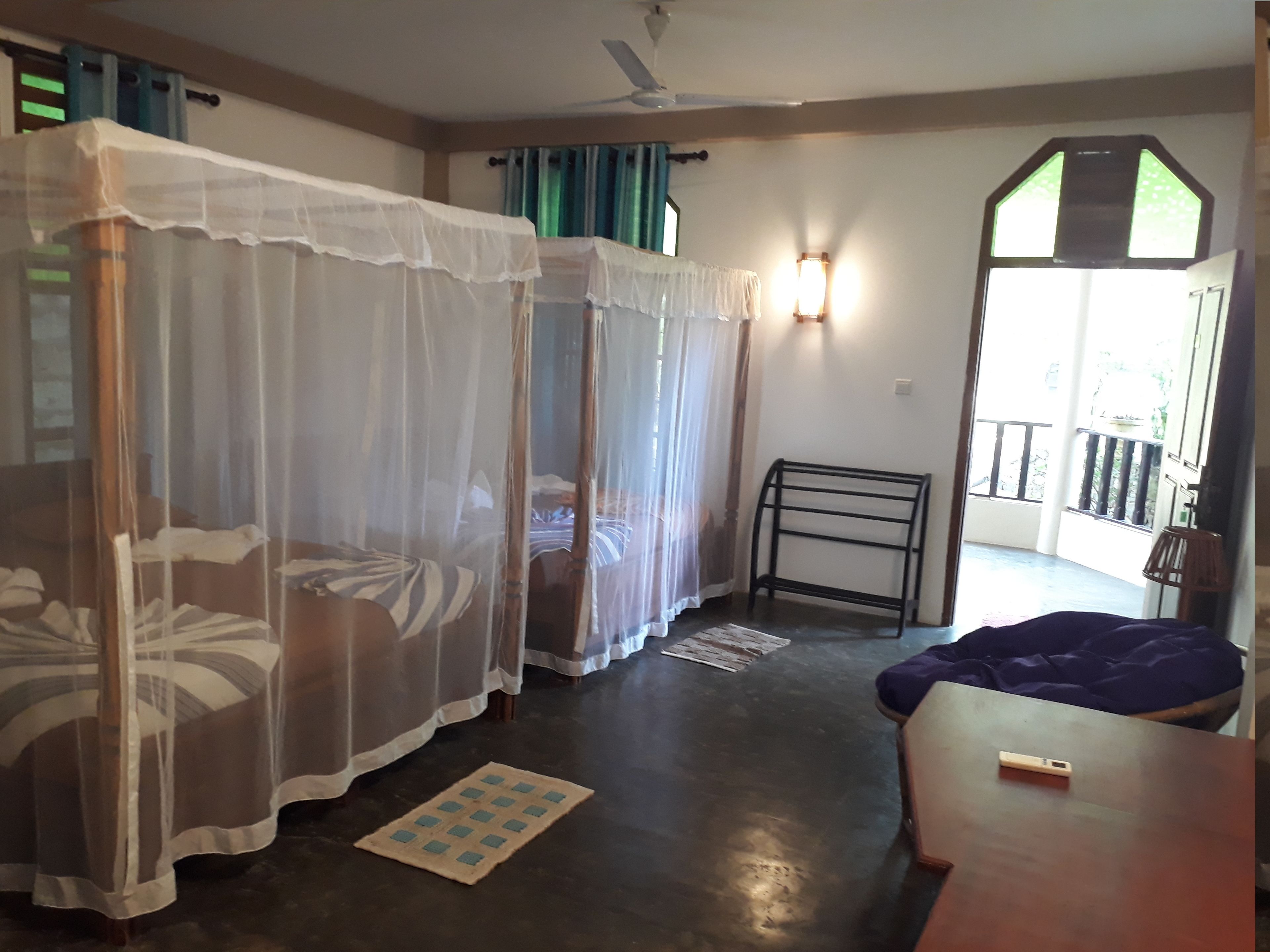 A room with four beds covered by mosquito nets, a small table, a bench, and a wall-mounted light fixture.