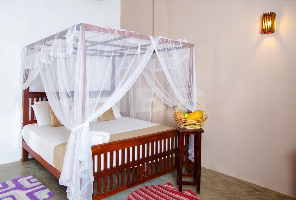 Een bed met houten frame en witte beddengoed, een witte hemel, een tafeltje met een mand vol fruit, en een wandlamp.