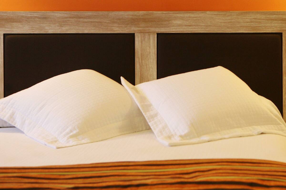 Een net opgemaakt bed met witte kussens en een gestreept dekbed, een houten hoofdbord, een klein houten nachtkastje met lade, en een oranje muur met een stopcontact.