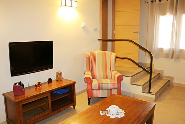 Una sala de estar con un televisor de pantalla plana, un sillón a rayas, una mesa de centro de madera y una escalera con barandilla.
