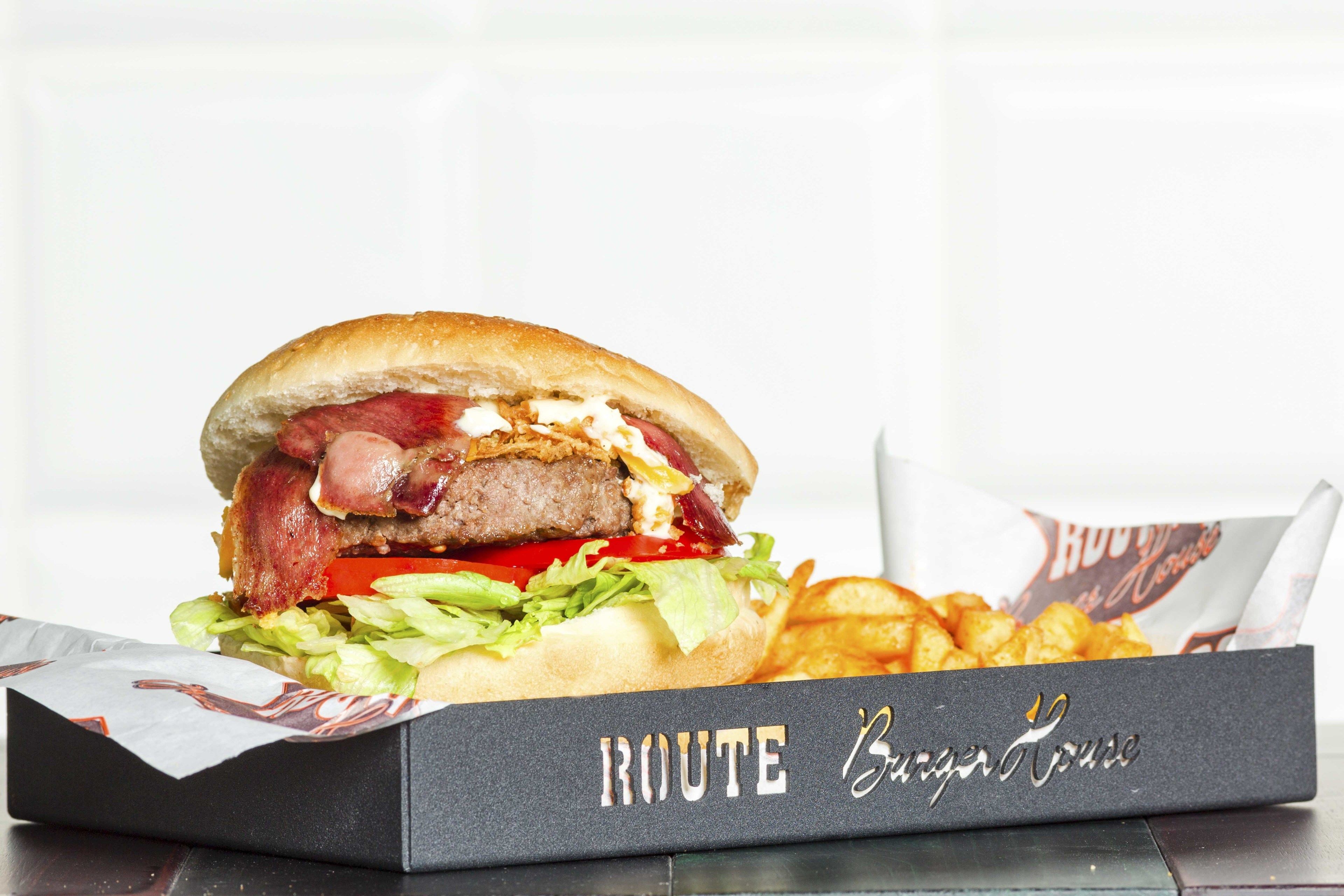 A burger with bacon, lettuce, tomato, and a bun, served with fries in a box labeled "ROUTE Burger House".