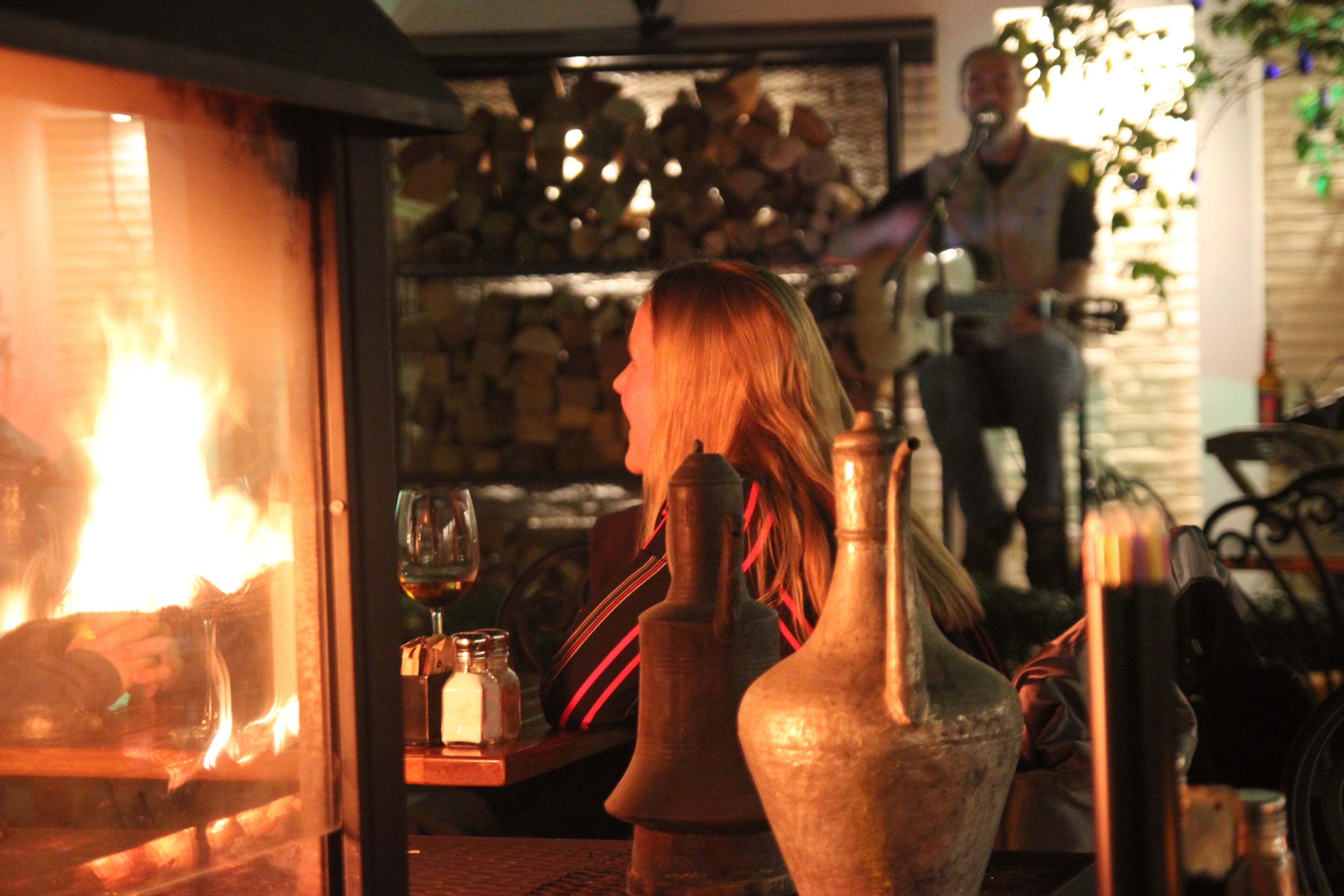 Una mujer sentada junto a la chimenea, con una copa de vino y saleros sobre una mesa.