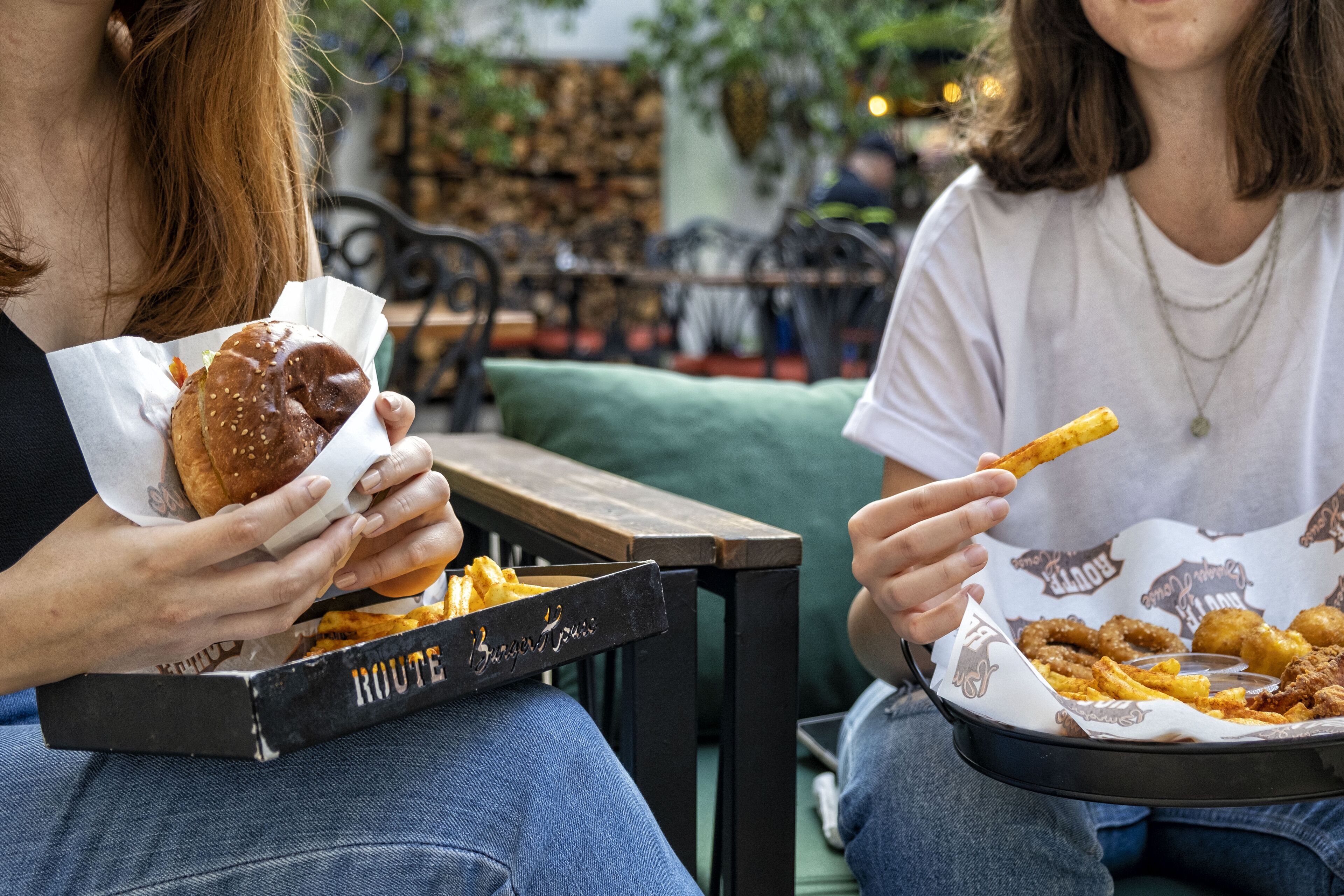 Duas pessoas sentadas à mesa com uma bandeja de comidas, incluindo um hambúrguer e batatas fritas.