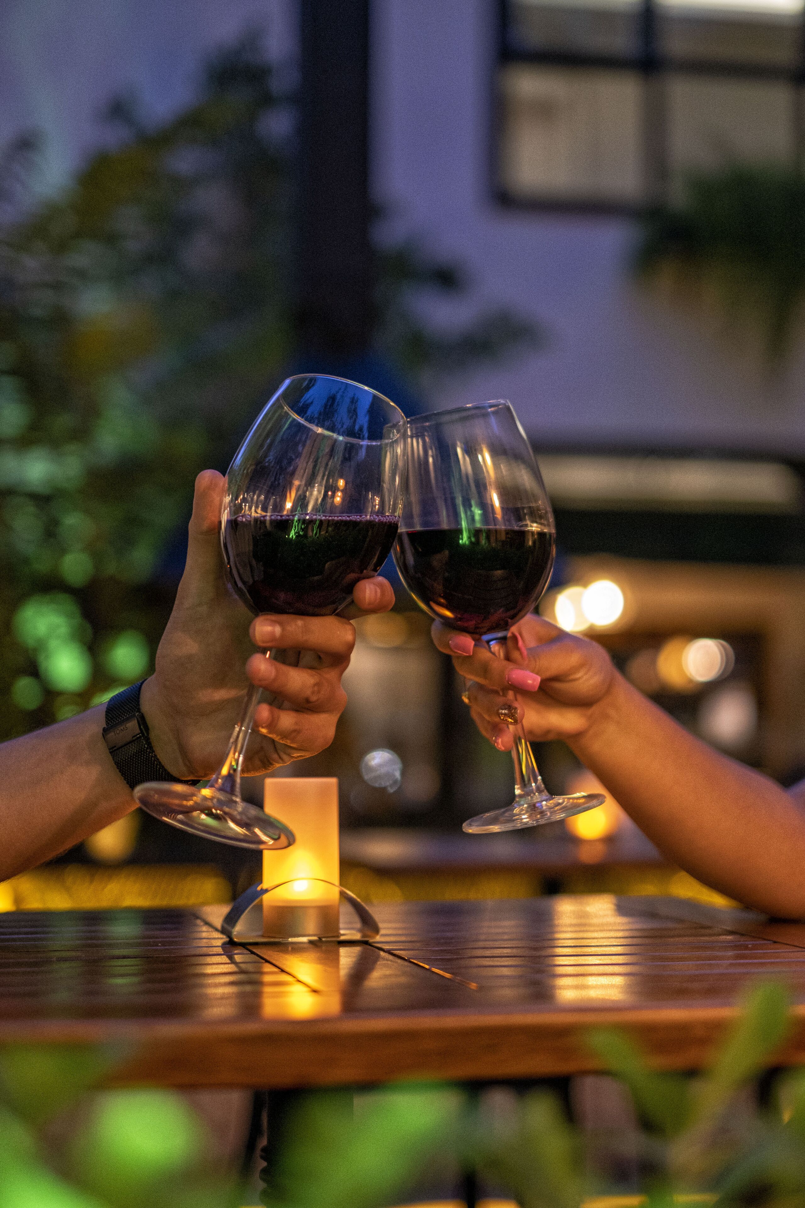 Two hands holding wine glasses at a table with a candle in the center.