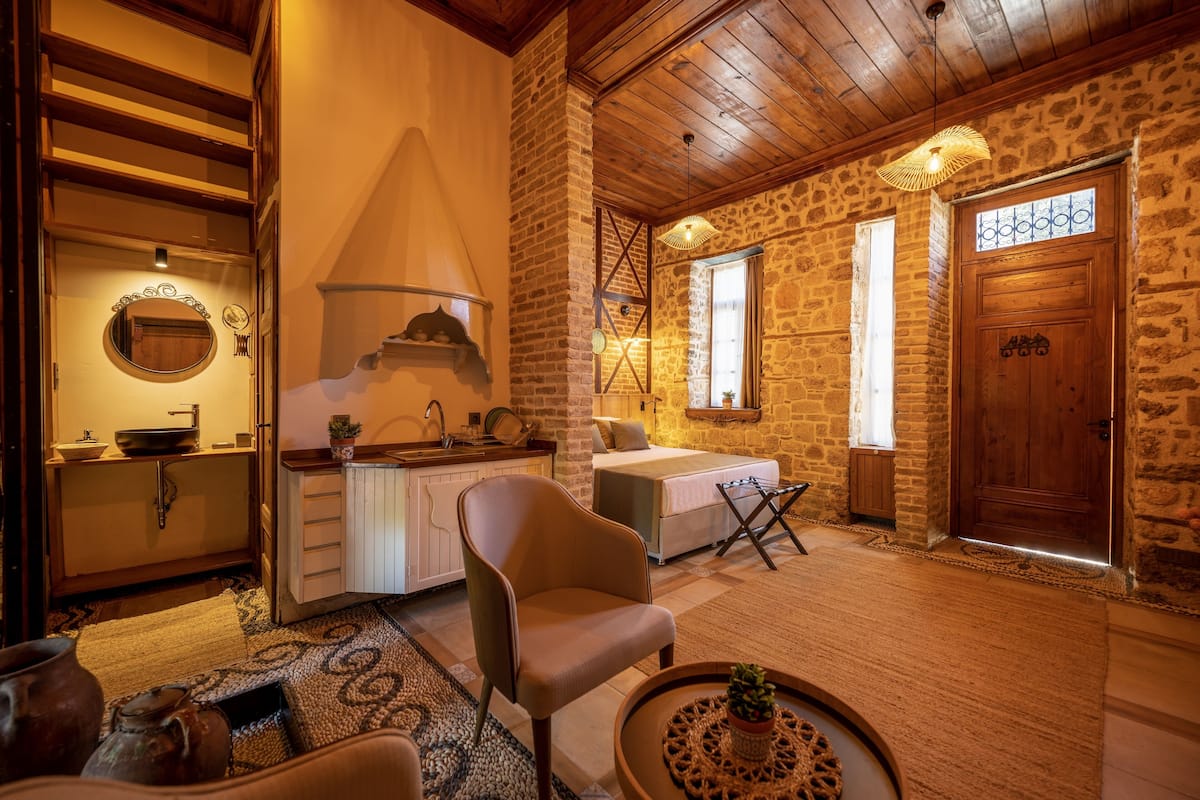 A cozy room with a stone wall, wooden ceiling, a bed, a chair, a small table, and a sink.