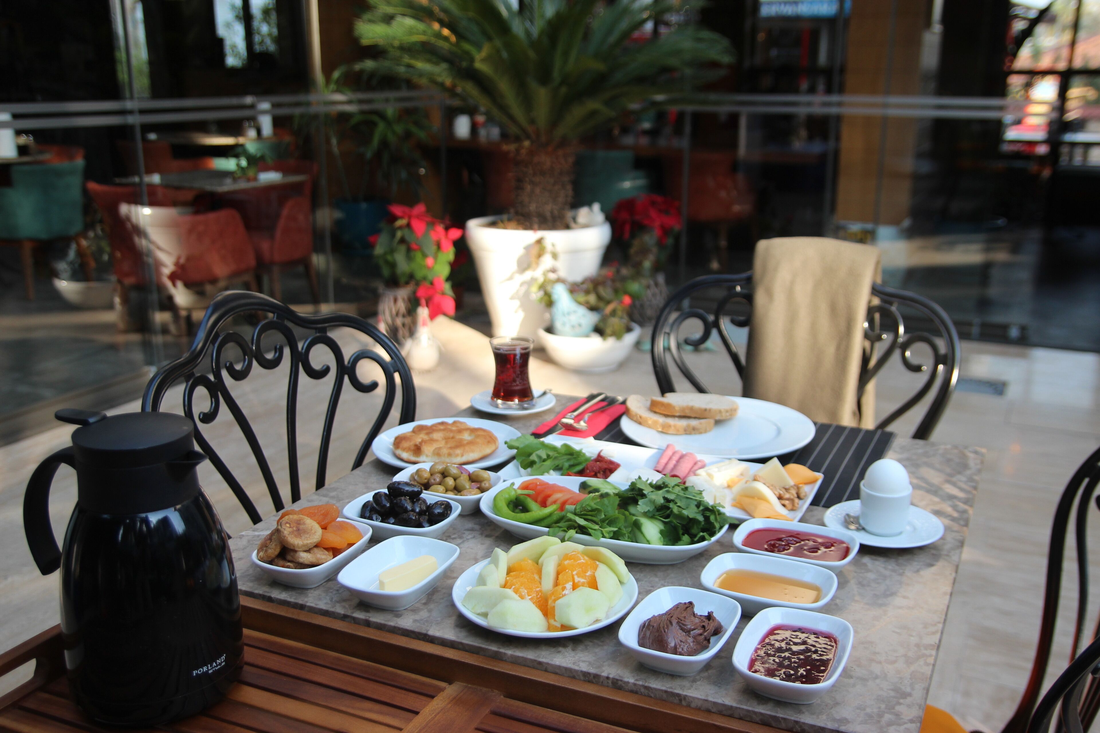 A table set with various dishes and a coffee pot, surrounded by chairs and potted plants.