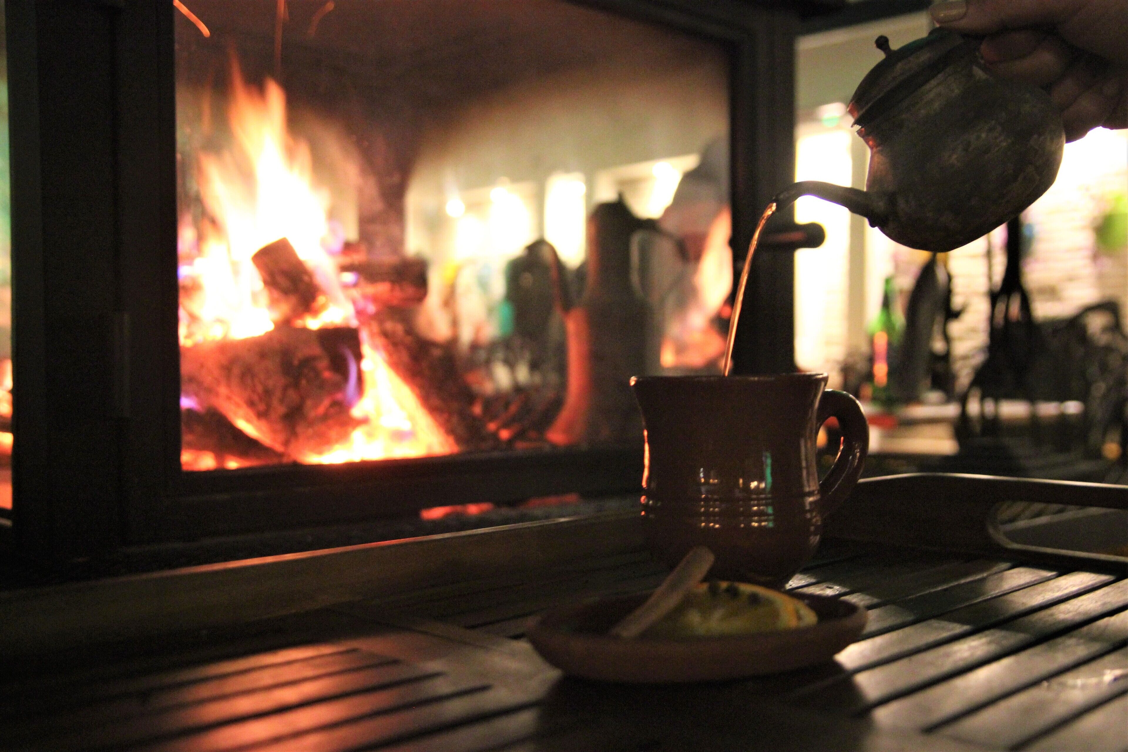 A mug on a table with a teapot pouring into it, in front of a fireplace.