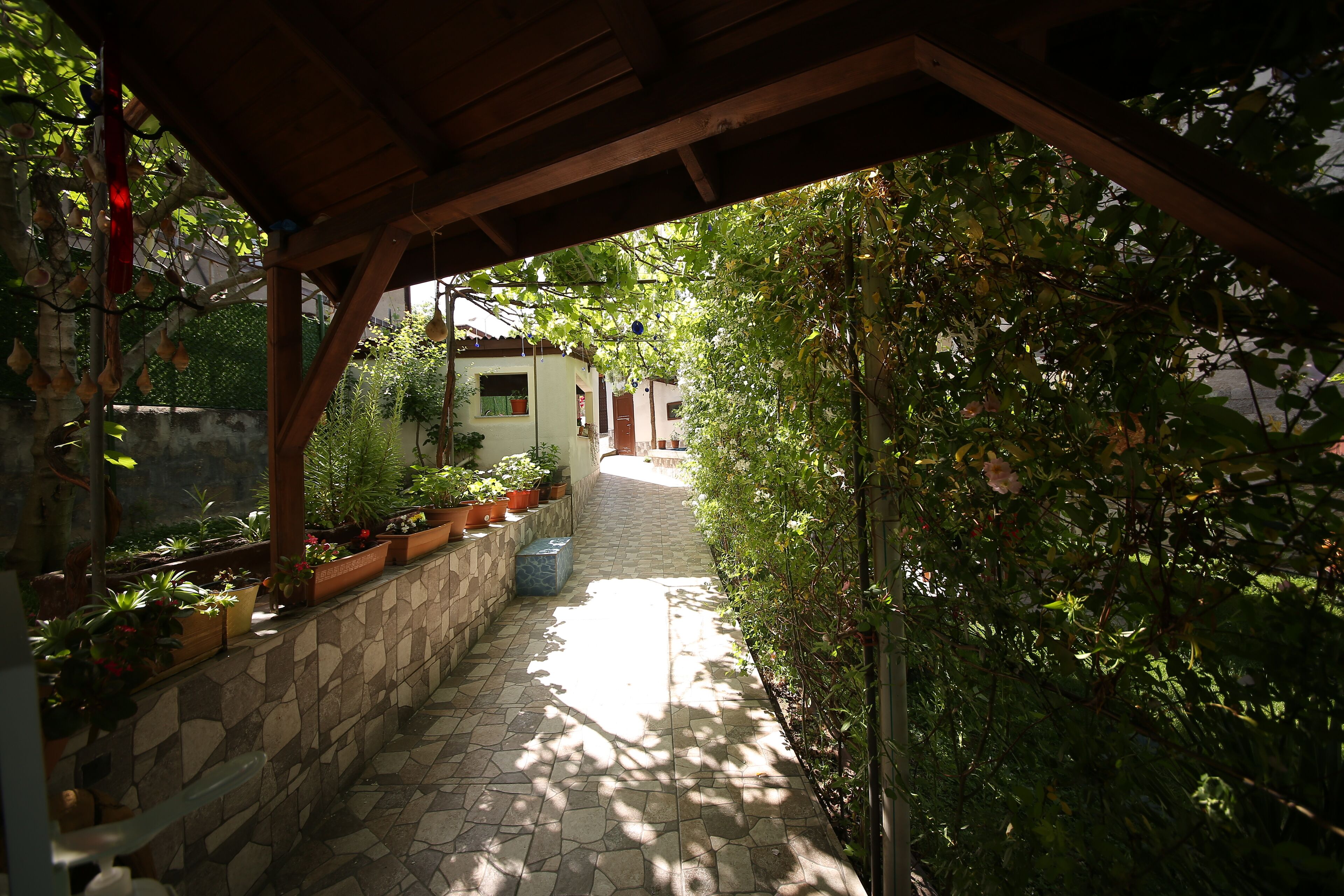 A stone-paved pathway leading to a covered area with wooden beams, surrounded by lush greenery and potted plants.
