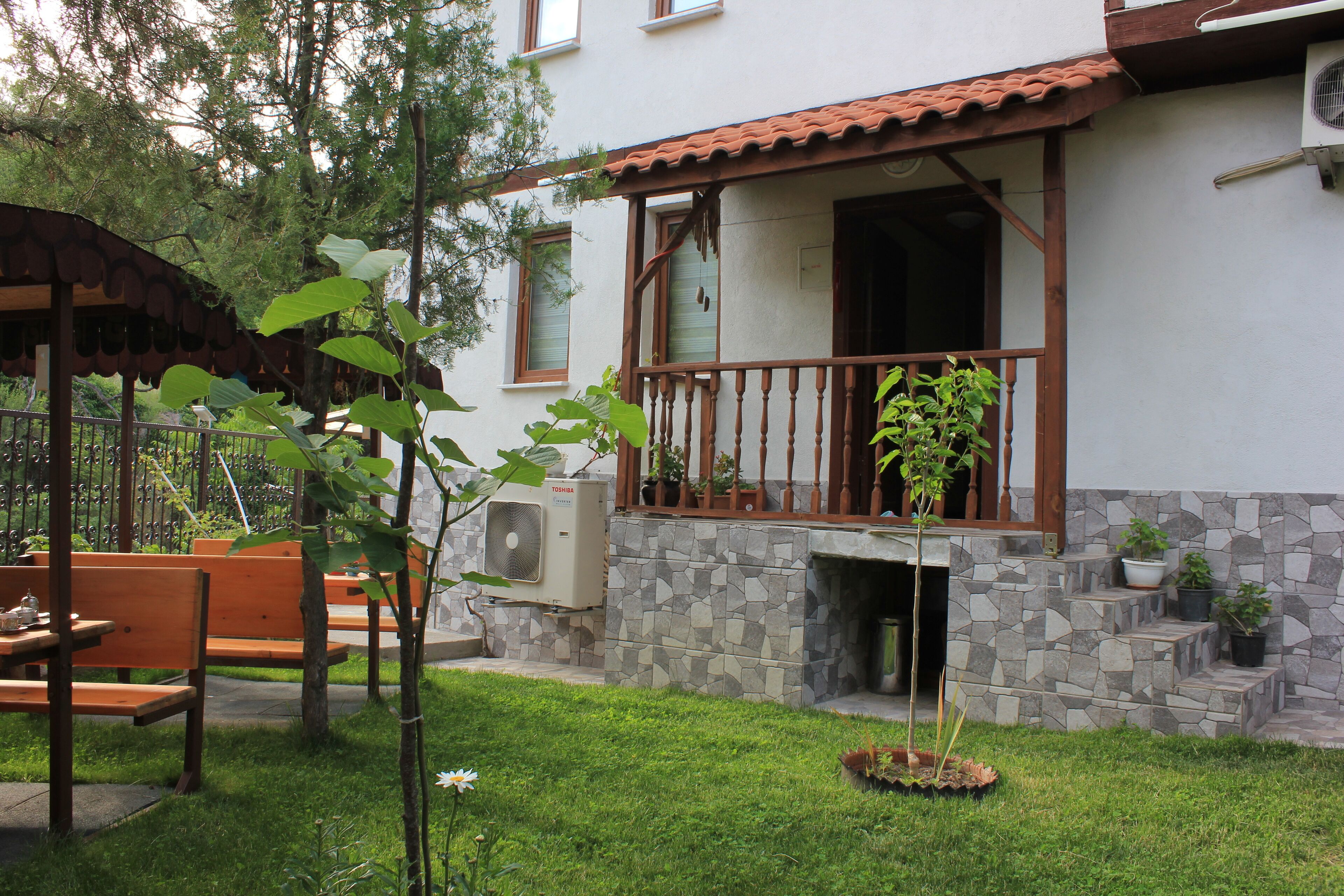 A residential building with a stone facade, a balcony with wooden railings, and a small garden area with benches and potted plants.
