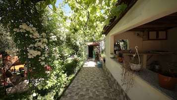 Um corredor estreito com piso de pedra e azulejos, ladeado por vegetação exuberante e plantas com flores brancas.