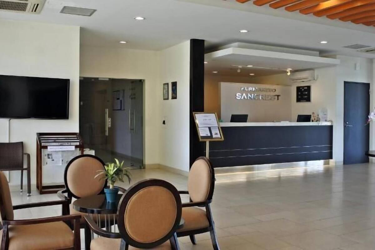 Uma área de recepção de hotel, com balcão de atendimento, televisão e um espaço de estar com cadeiras e mesa.