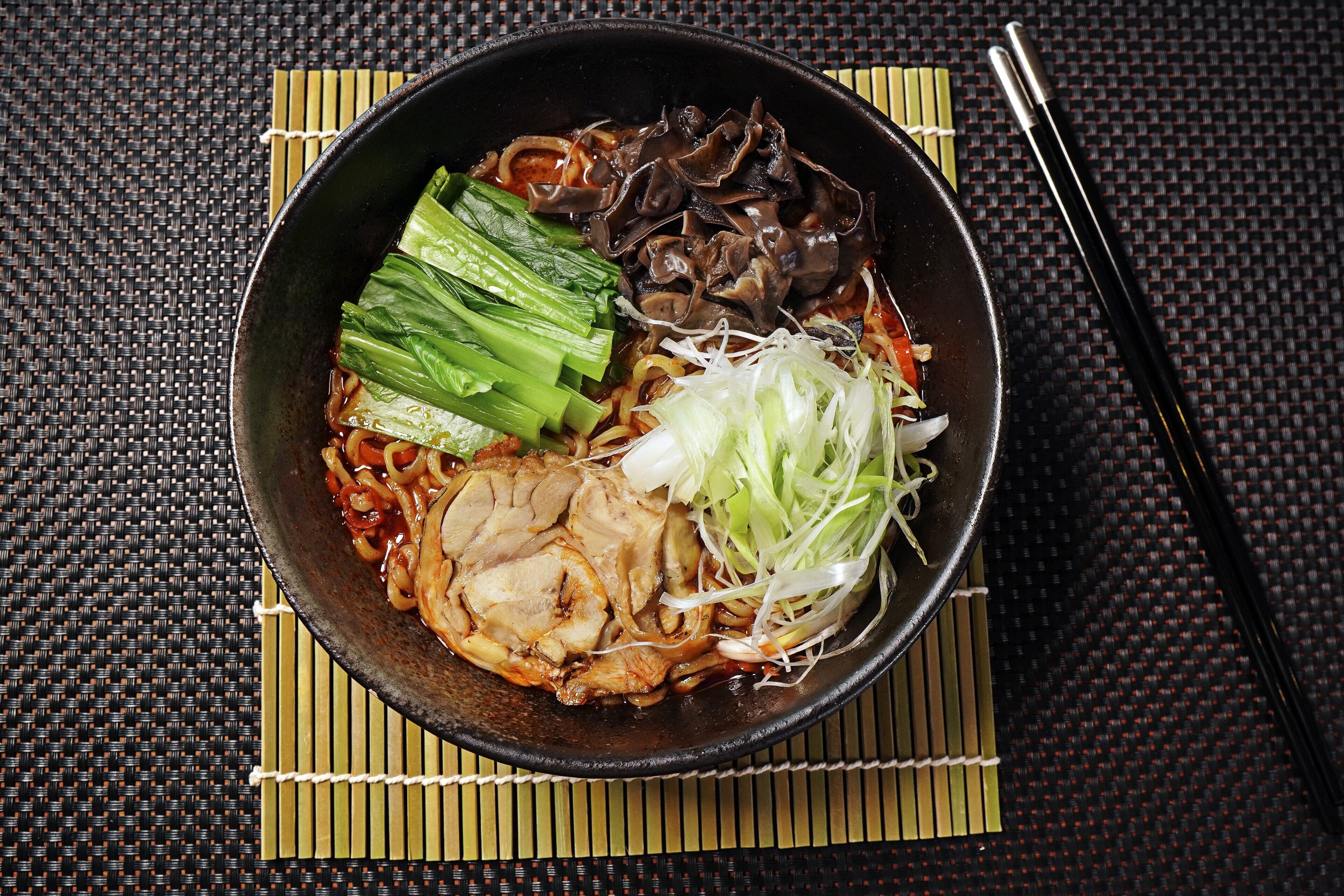 Eine Schüssel Ramen mit verschiedenen Belägen, darunter Bok Choy und Pilze, auf einer gewebten Matte abgestellt.