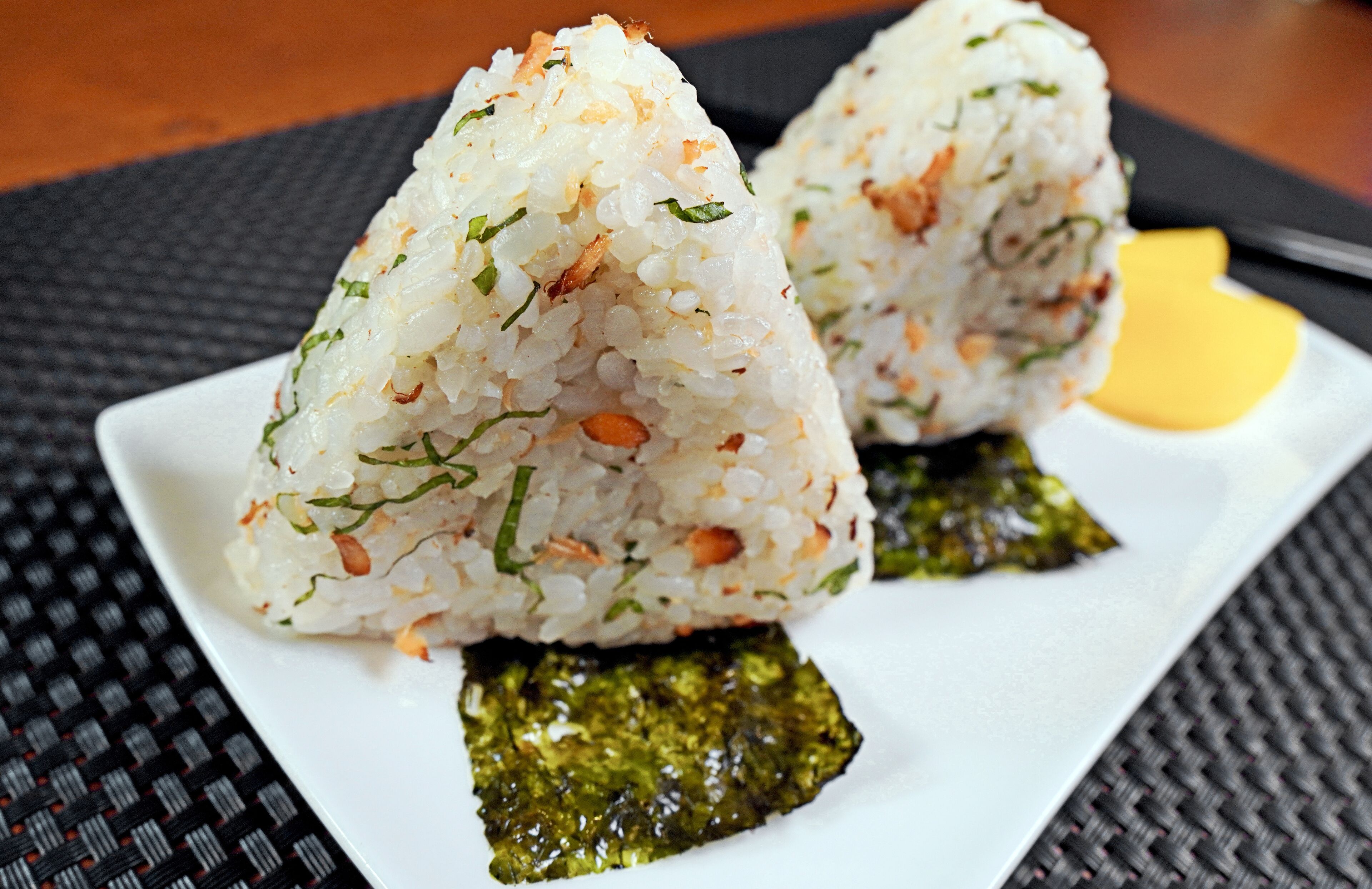 Une assiette contenant deux portions de riz en forme de triangle, un morceau d’algue et un objet jaune, le tout posé sur une table noire à texture marquée.