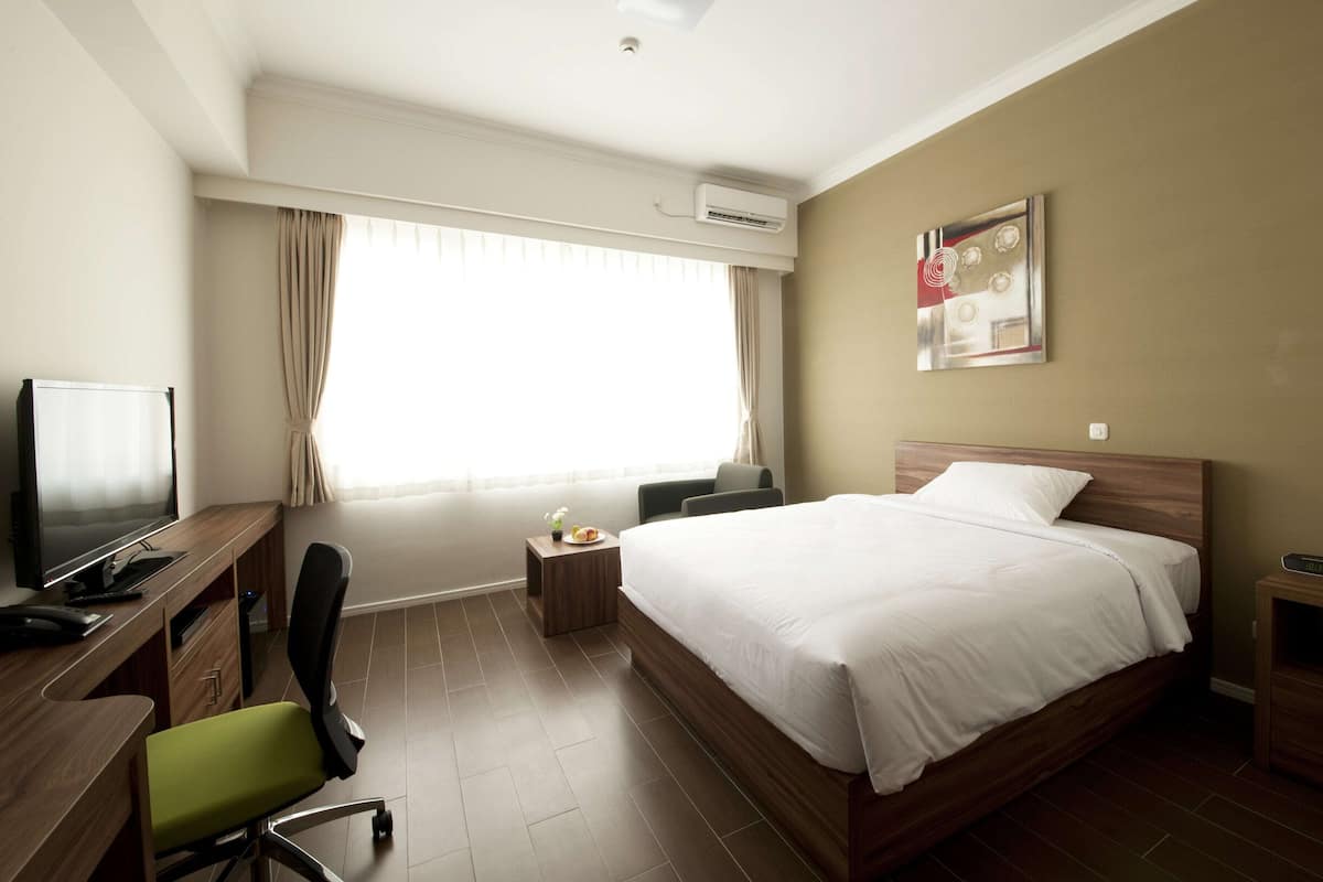 Quarto de hotel com cama, escrivaninha com computador, cadeira, pequena mesa com planta e janela com cortinas.