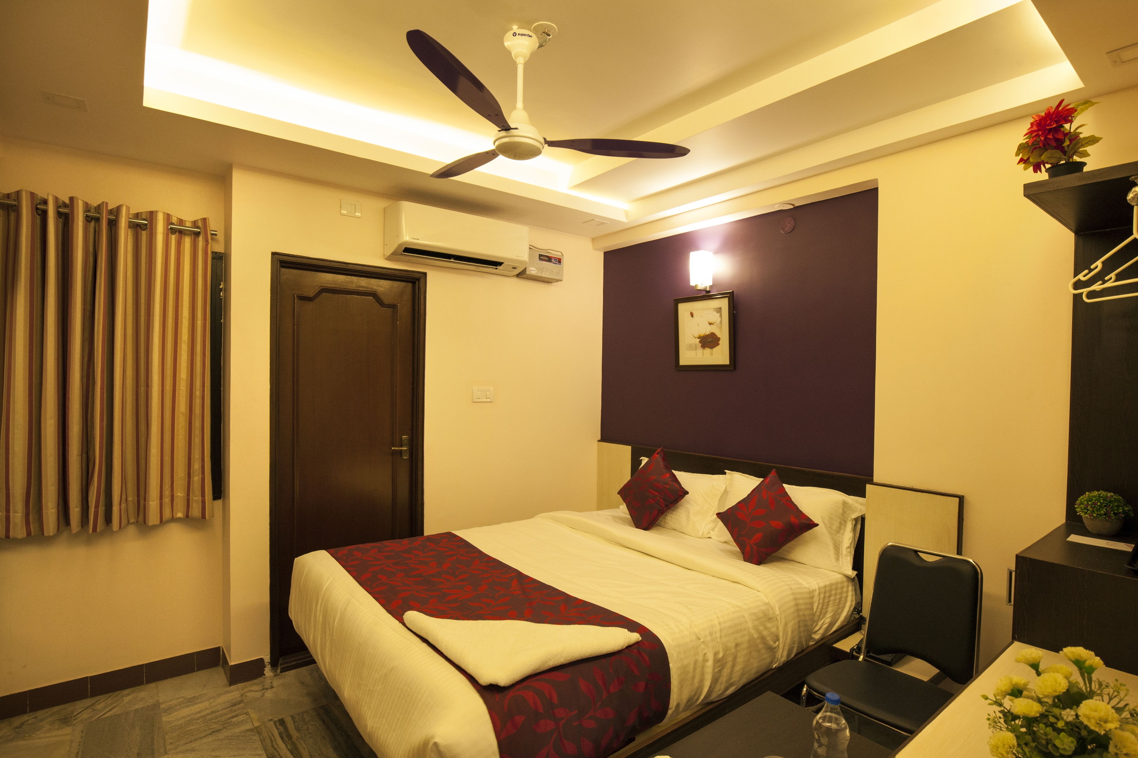 A hotel room with a bed, a ceiling fan, an air conditioning unit, a desk with a chair, and a small table with a vase of flowers.