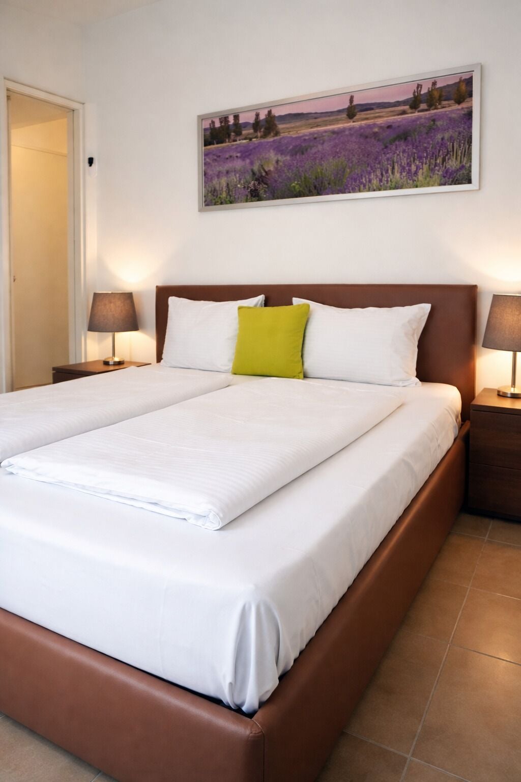 A neatly made bed with white linens and a brown headboard, flanked by two bedside tables with lamps, in a room with a large landscape painting above the bed.