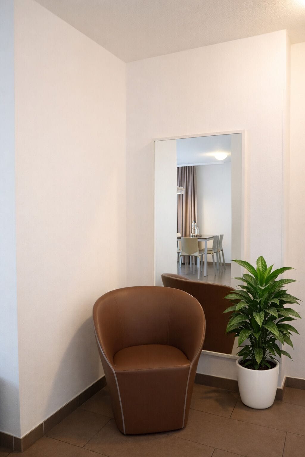 Een hoek van een hotelkamer met een bruine stoel, een plant in pot en een spiegel die een zithoek weerkaatst.