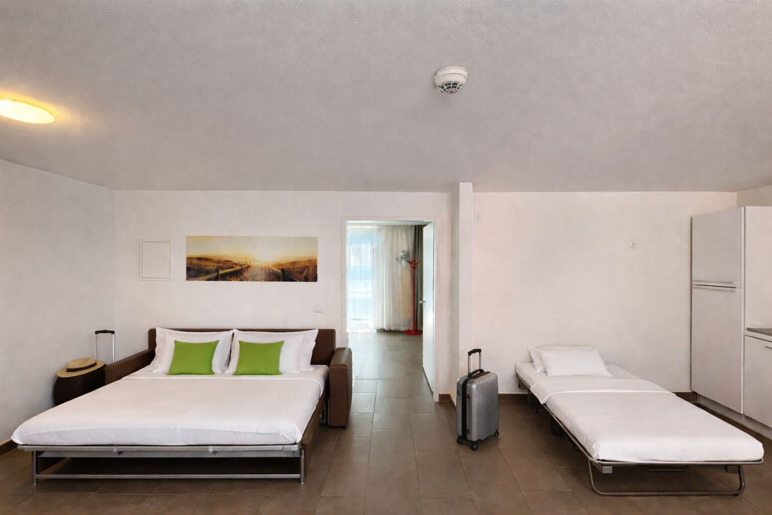 A hotel room with a large bed, a smaller bed, a suitcase, a wardrobe, and a painting on the wall.