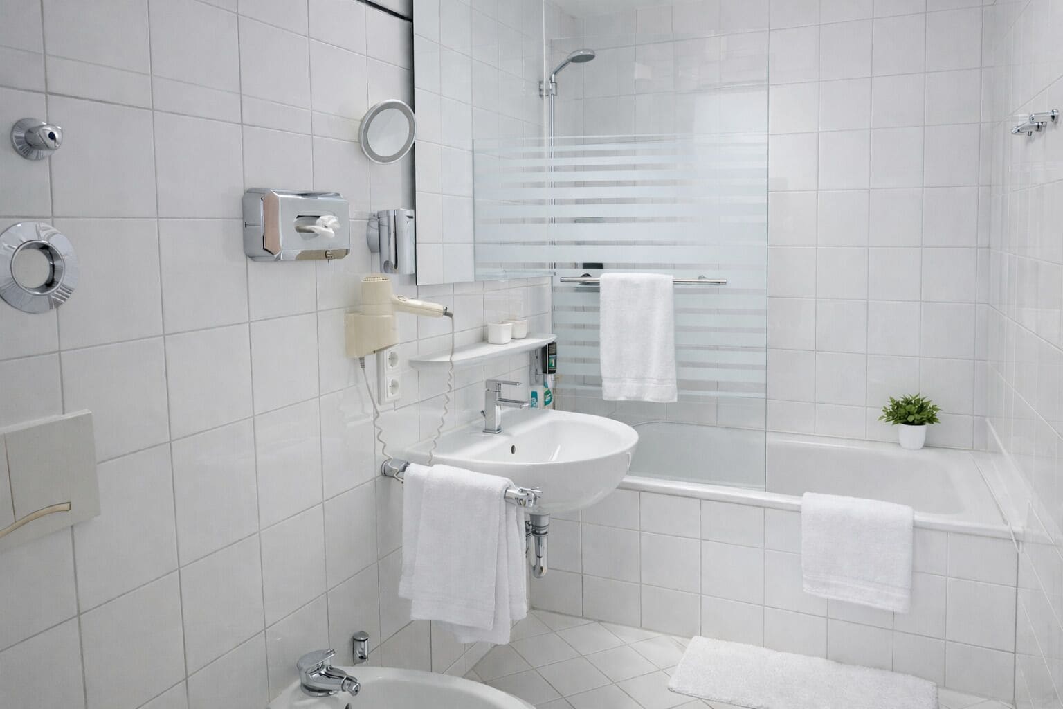 A modern bathroom with a white sink, mirror, and shower. There are towels hanging on the wall and a small plant on the floor.