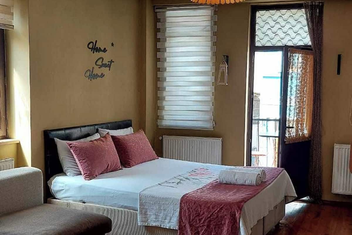 A bedroom with a bed, a sofa, a window with blinds, a hanging lamp, and a wall with a decorative phrase.