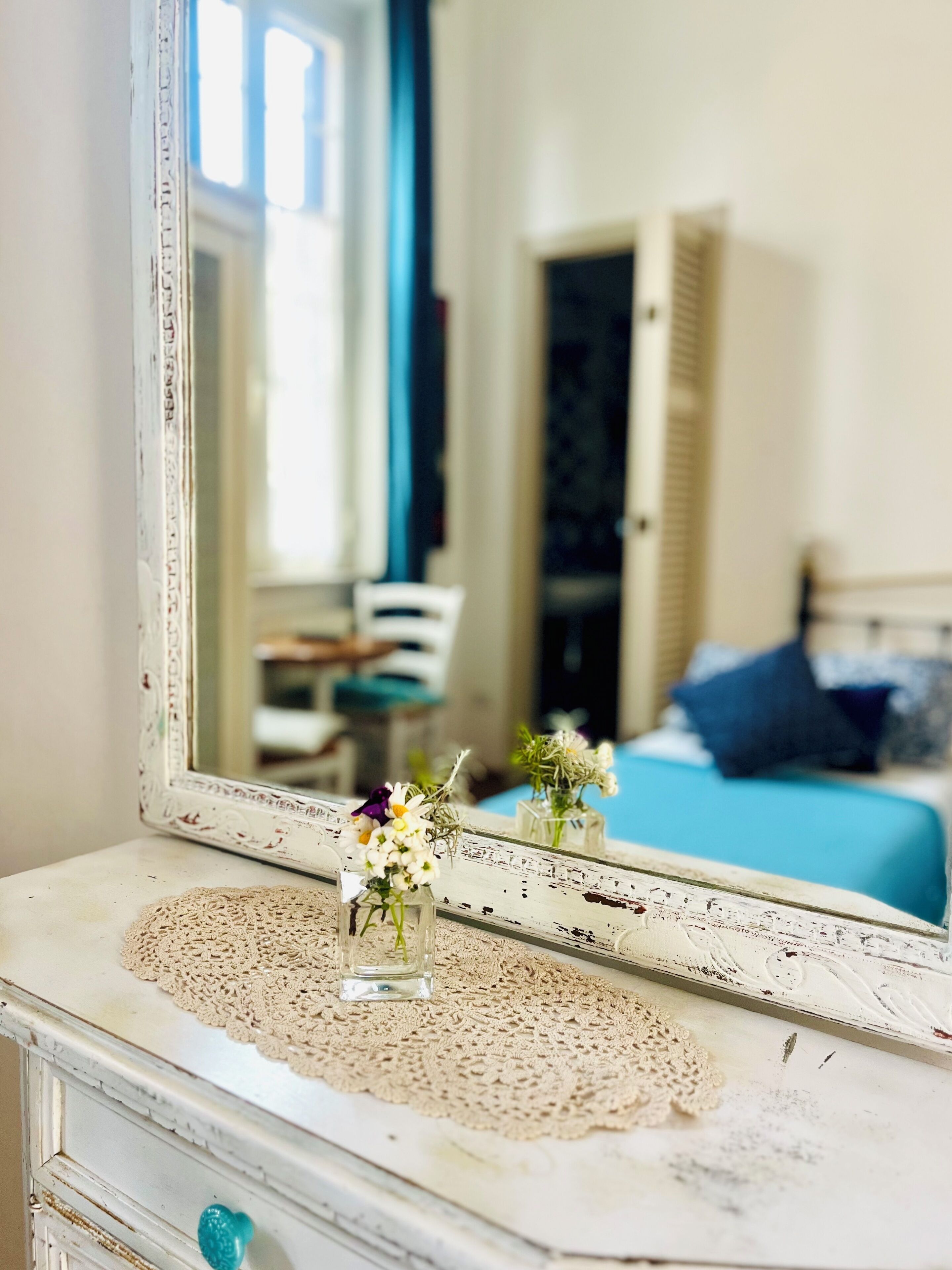 A mirror reflecting a room with a bed, a chair, and a dresser with a lace doily.
