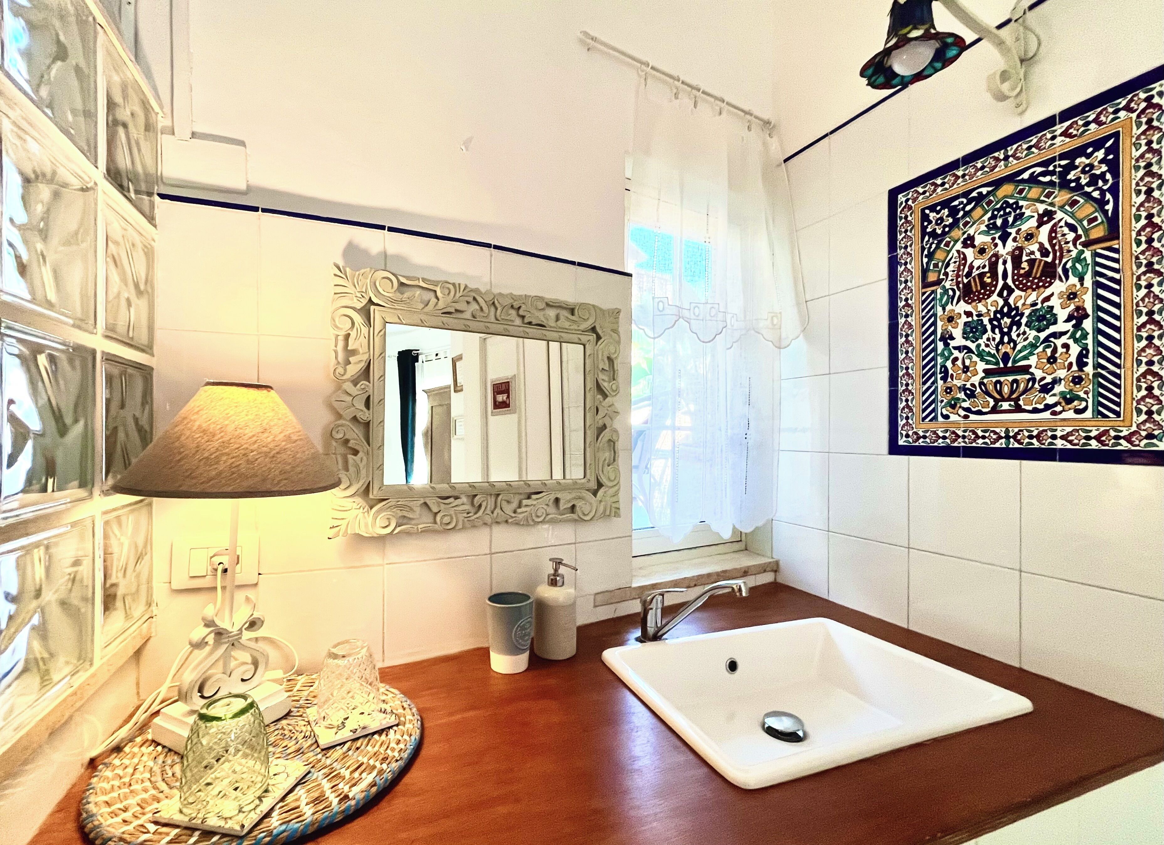 A bathroom with a white sink, a mirror with an ornate frame, a wall-mounted lamp, and a decorative tile with floral patterns.
