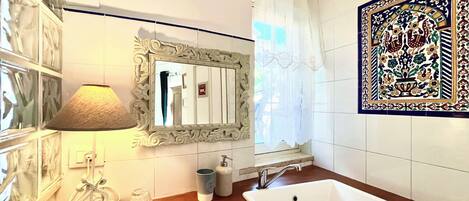 A bathroom with a white sink, a mirror with an ornate frame, a wall-mounted lamp, and a decorative tile with floral patterns.