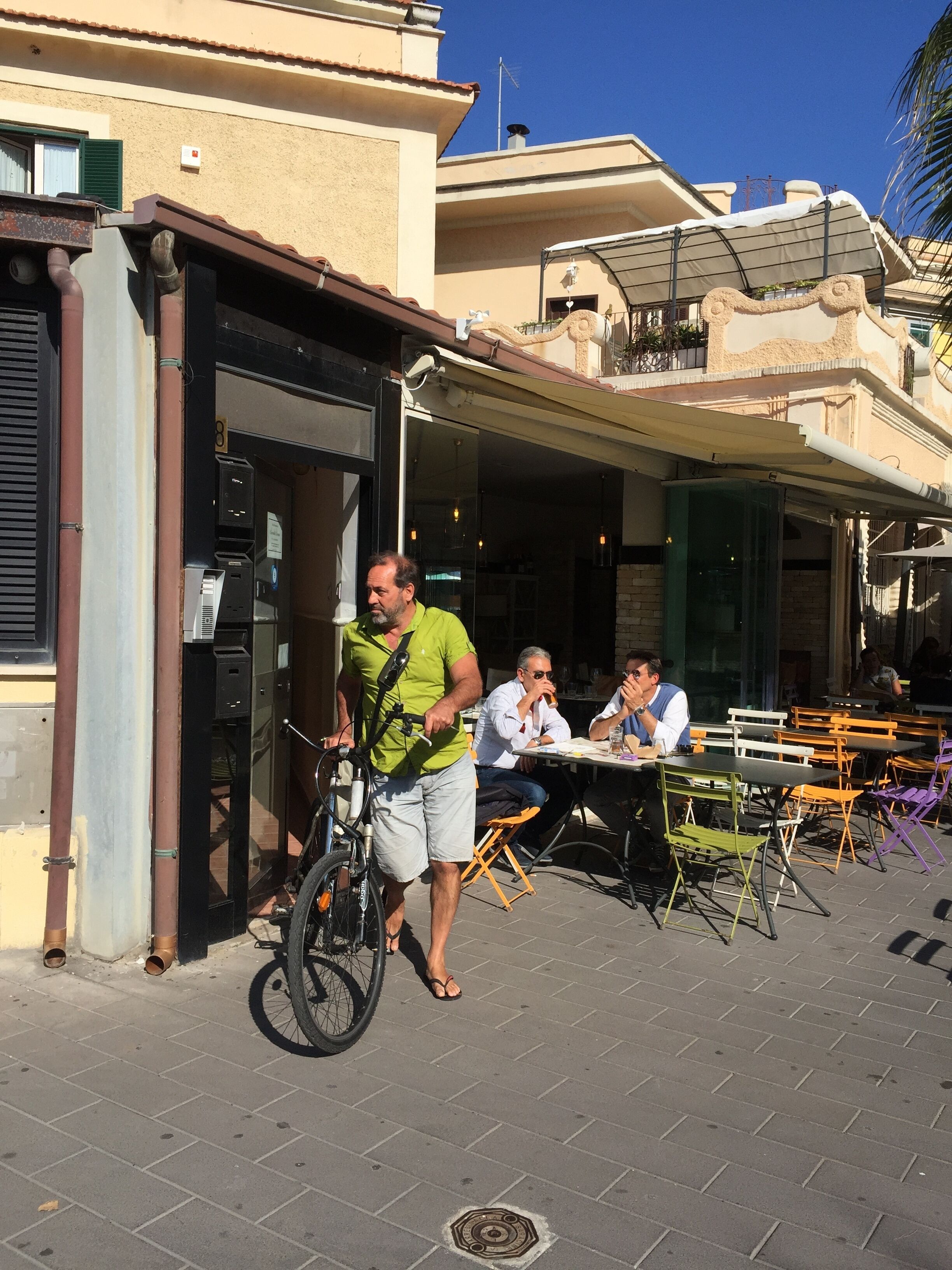 Um café ao ar livre com mesas e cadeiras, uma pessoa com bicicleta e um edifício com telhado em azulejos.