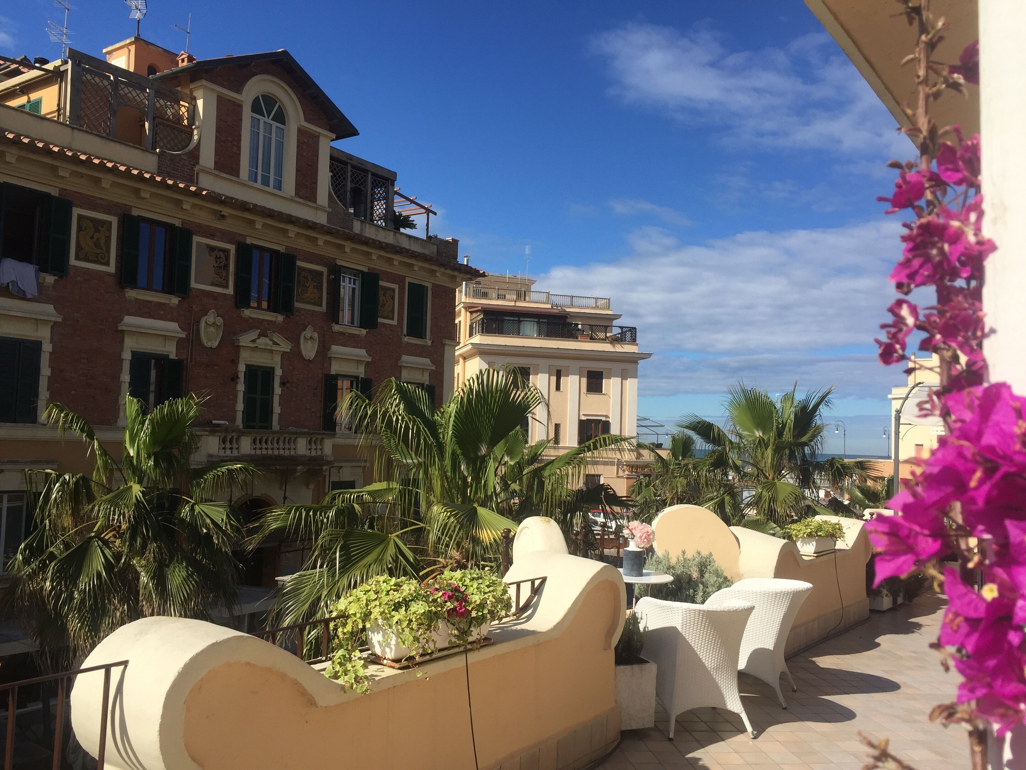 A hotel balcony with outdoor seating, palm trees, and a view of the sea.