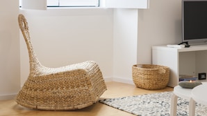 Een kamer met een geweven stoel, een klein rond tafeltje, een vloerkleed met patroon, een televisie op een witte standaard en een raam met jaloezieën.