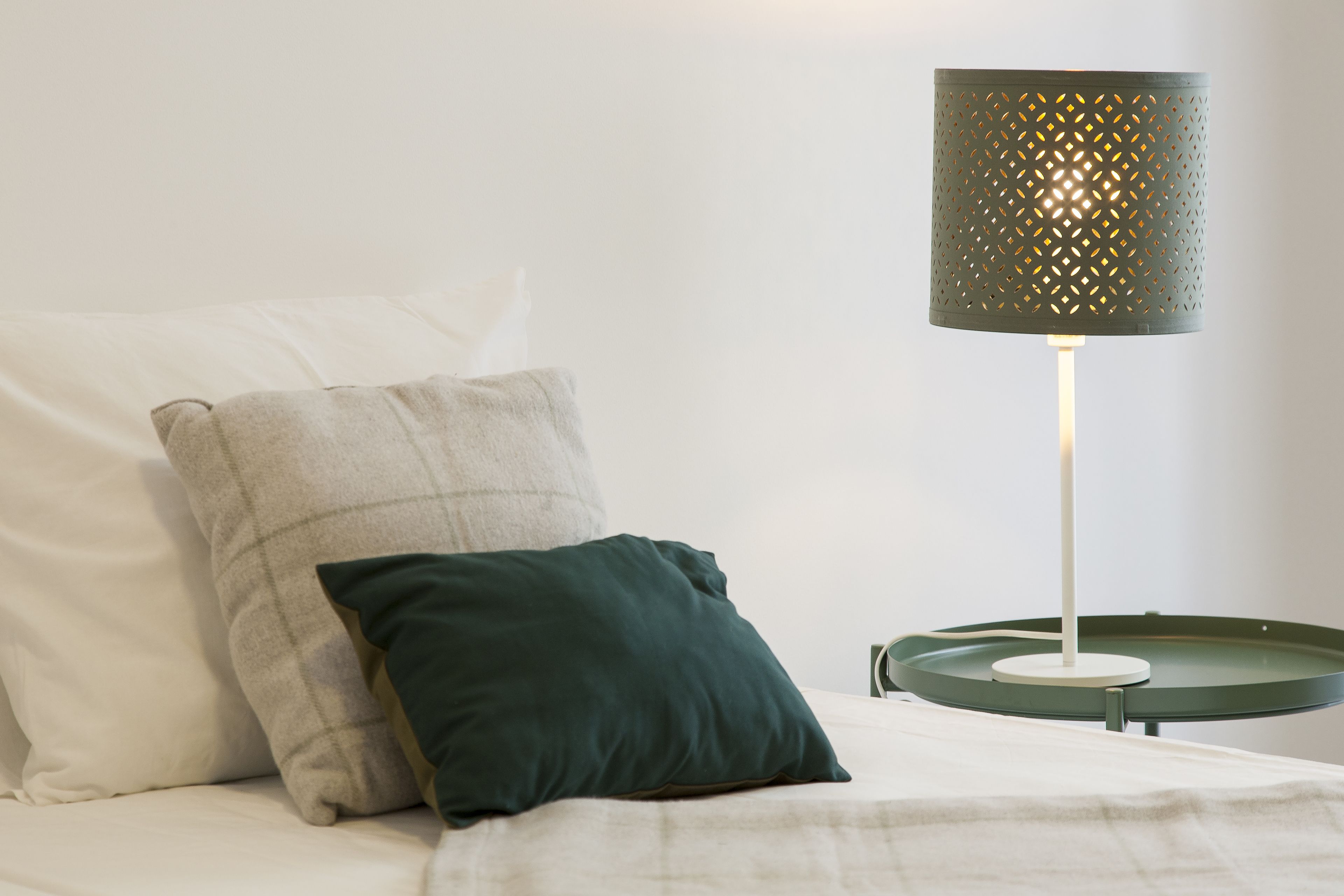 Una cama con dos almohadas, una lámpara con pantalla perforada y una mesita de noche con una vela.