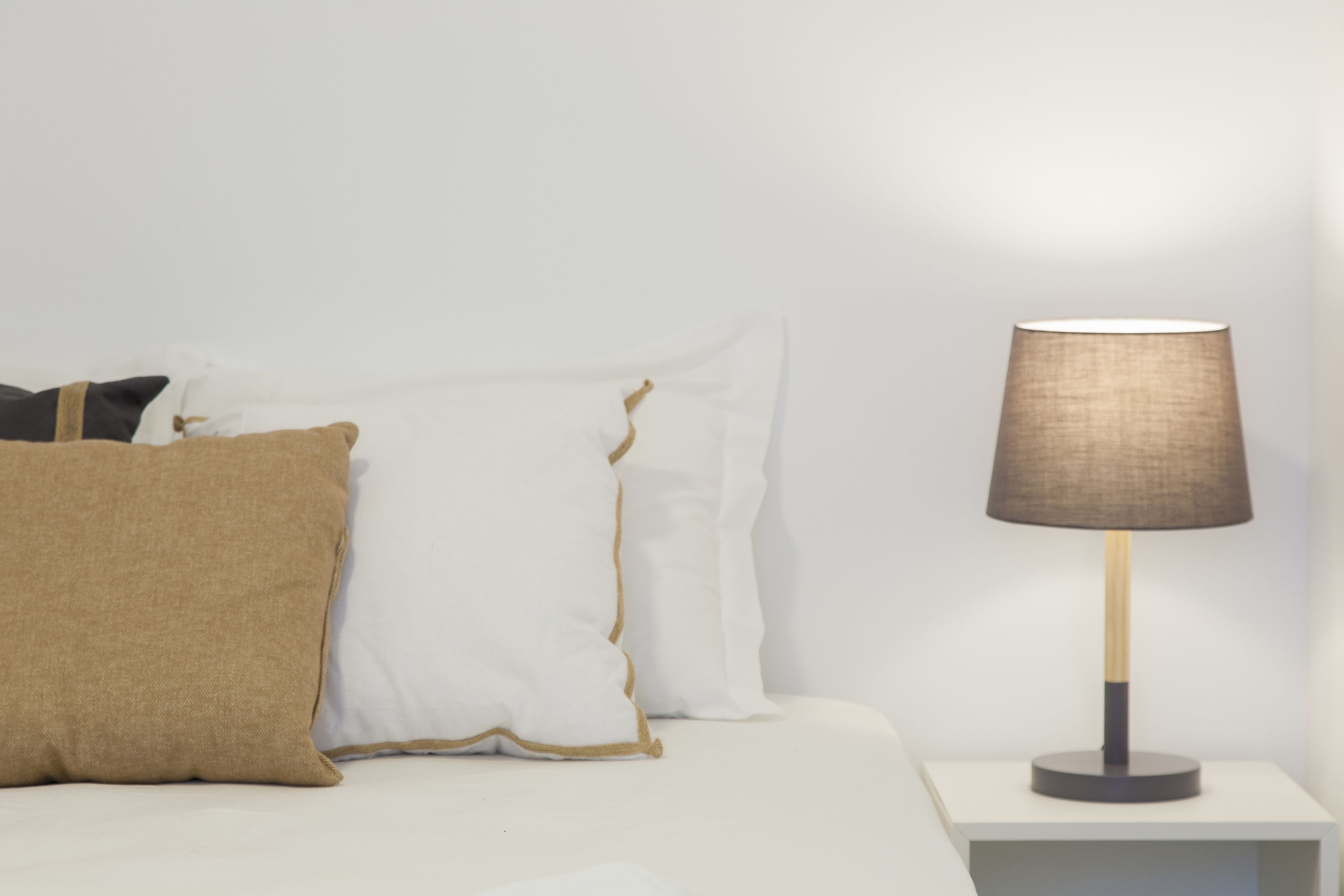Una cama con almohadas blancas y beiges, una mesita de noche con una lámpara y una pared blanca.