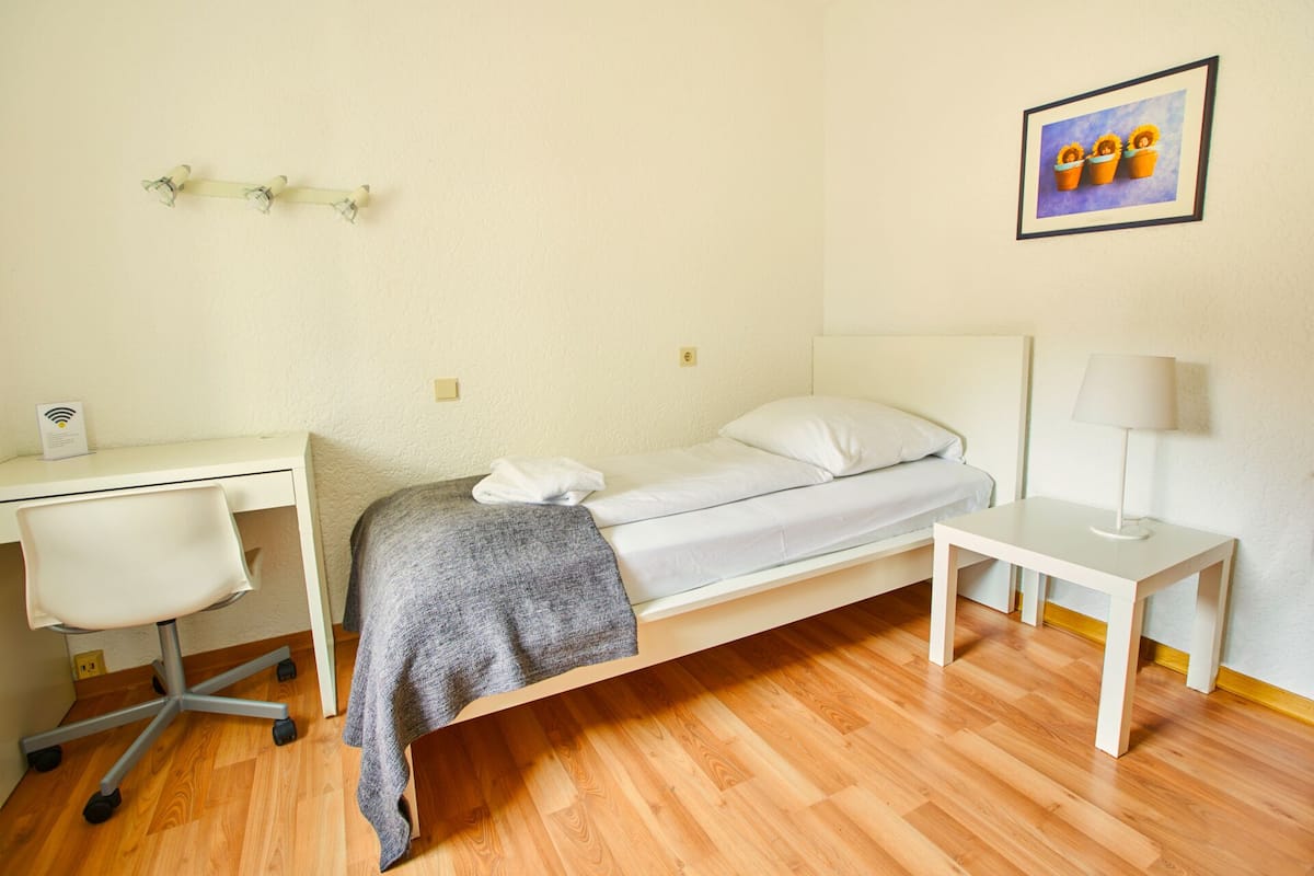A small, neatly arranged bedroom with a single bed, a desk, and a chair. There is a framed picture on the wall and a lamp on the desk.