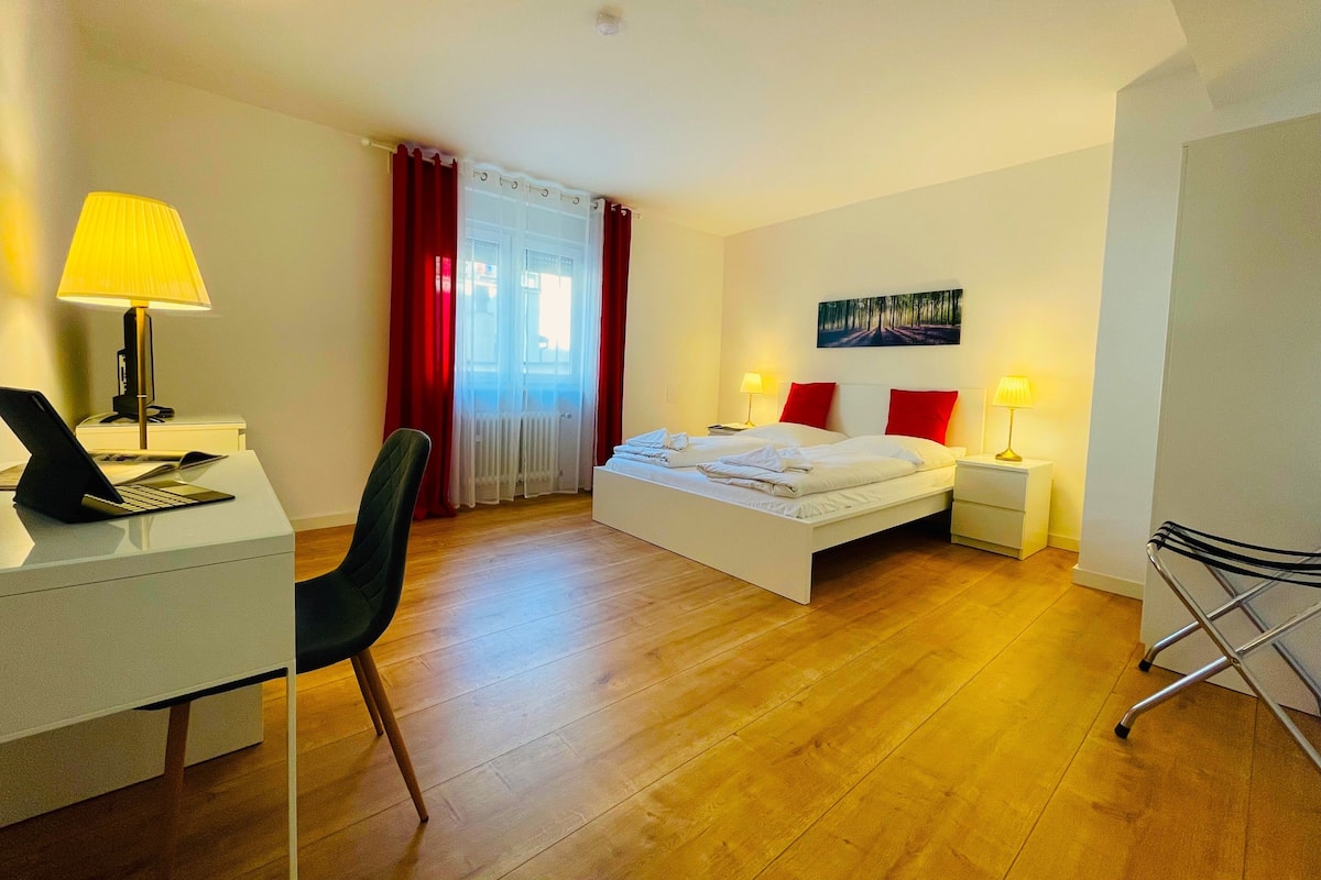 A hotel room with a bed, a desk with a laptop, a chair, a bedside table with a lamp, and a window with curtains.