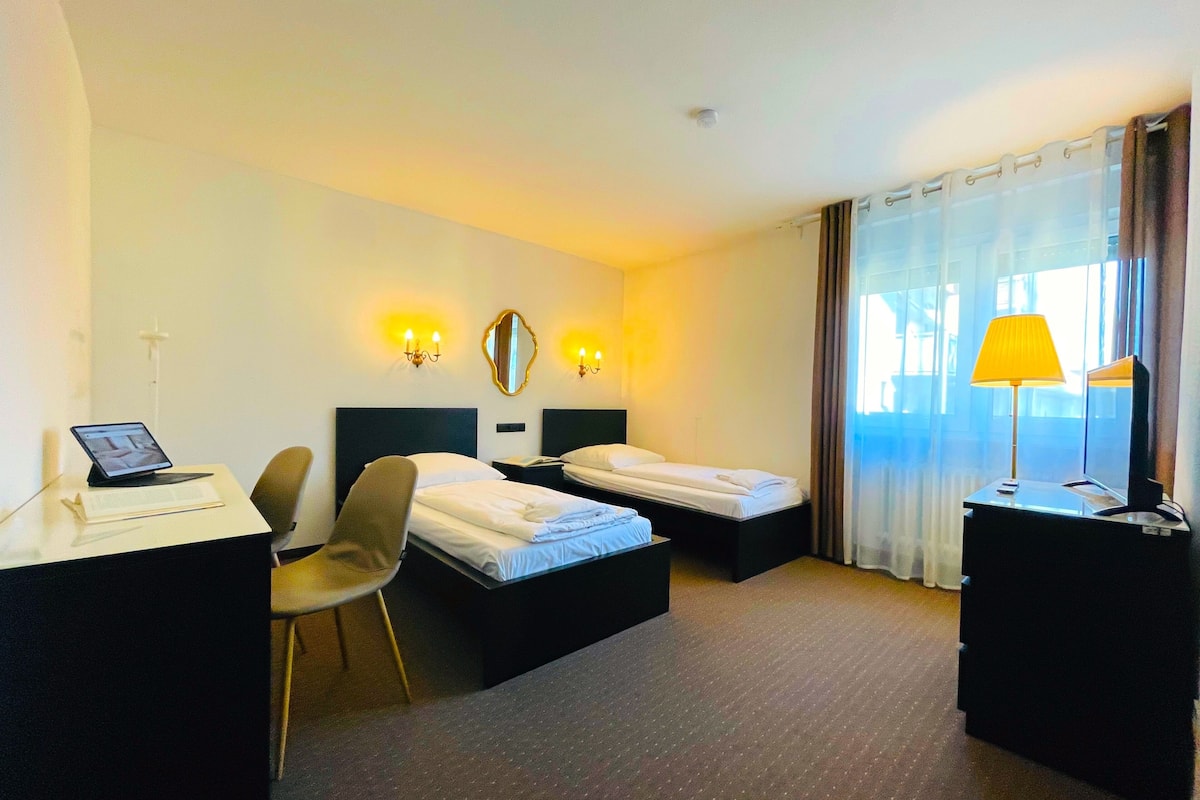 A hotel room with two beds, a desk with a laptop, a chair, a lamp, and a large window with curtains.