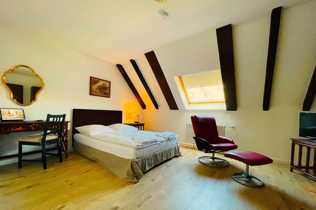 A bedroom with a bed, desk, chair, and a red chair. There is a window with a view of the outside.