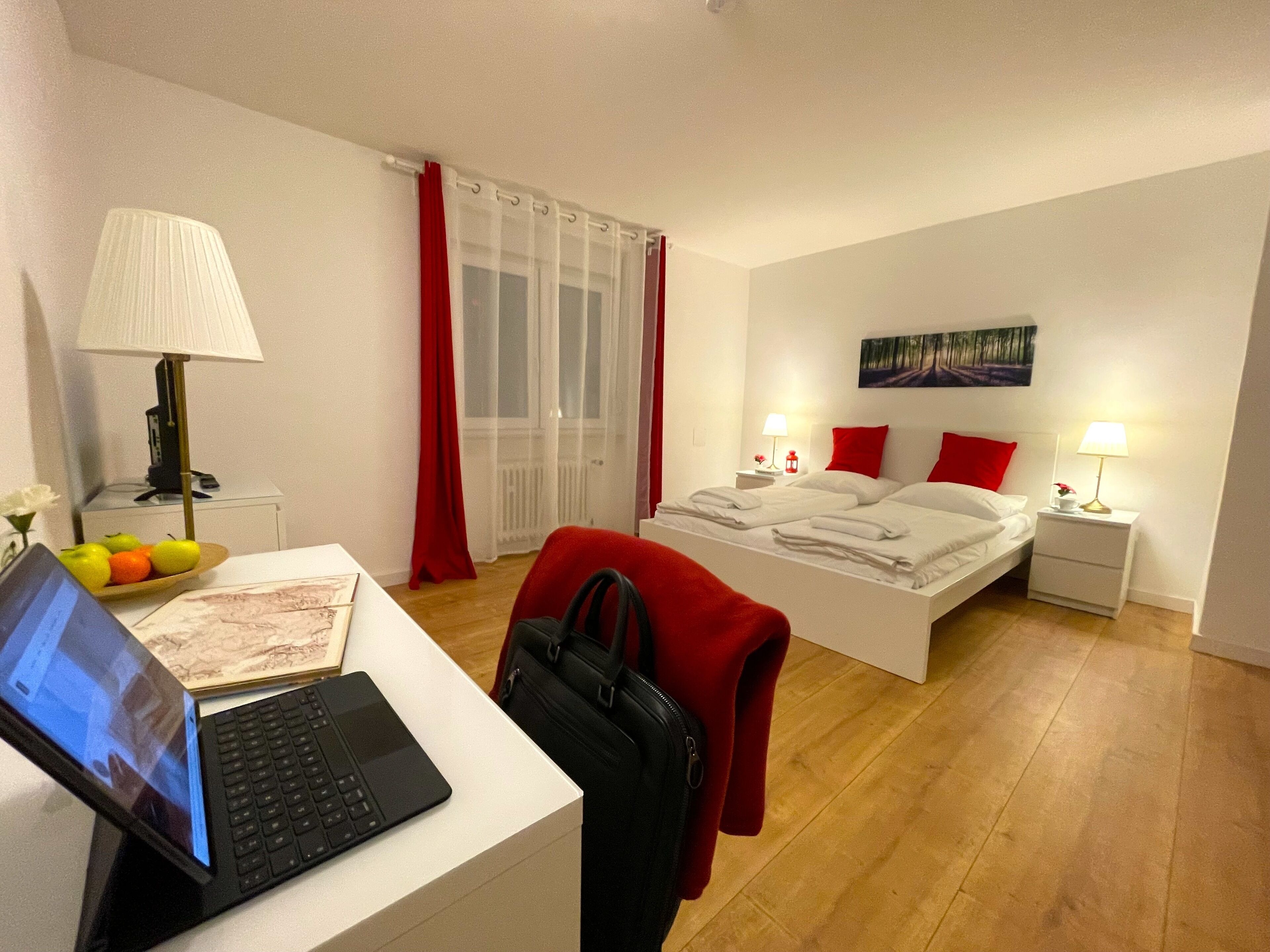 Ein Hotelzimmer mit Bett, Schreibtisch mit Laptop, Stuhl, Koffer, Nachttisch mit Lampe und Fenster mit Vorhängen.