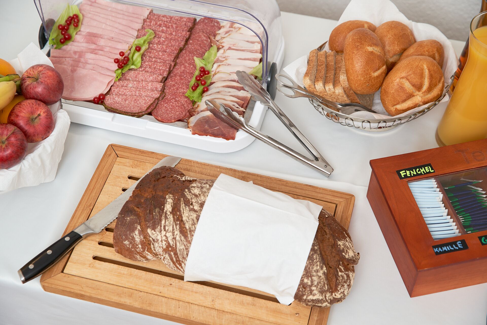 Un bufé de desayuno con jamón, salami, pan, manzanas y una tabla de cortar con cuchillo.