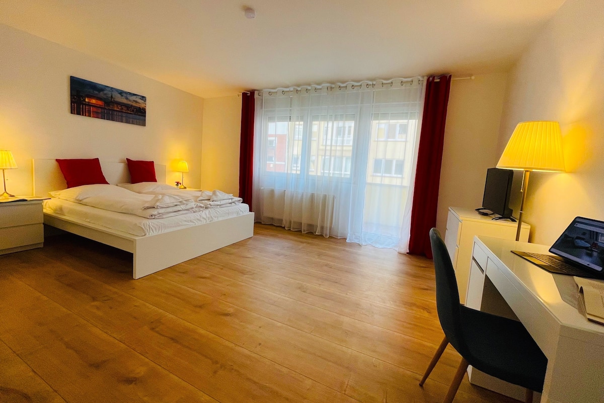 A hotel room with a bed, a desk, and a chair. There is a window with curtains and a painting on the wall.