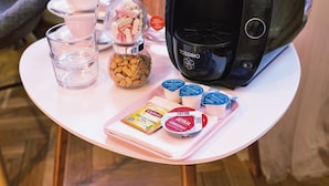 A table with a coffee maker, jars of nuts and candies, glasses of water, and packets of tea and coffee.