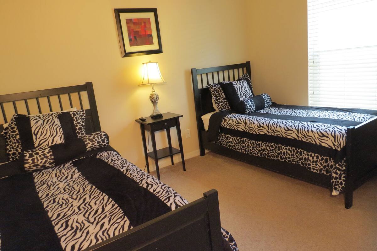 A bedroom with two beds, each with zebra-patterned bedding, a nightstand with a lamp, and a framed picture on the wall.