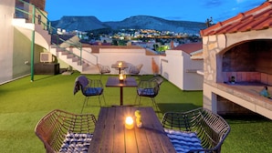 A terrace with a table and chairs, a barbecue area, and a view of a town and mountains in the background.