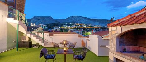 A terrace with a table and chairs, a barbecue area, and a view of a town and mountains in the background.