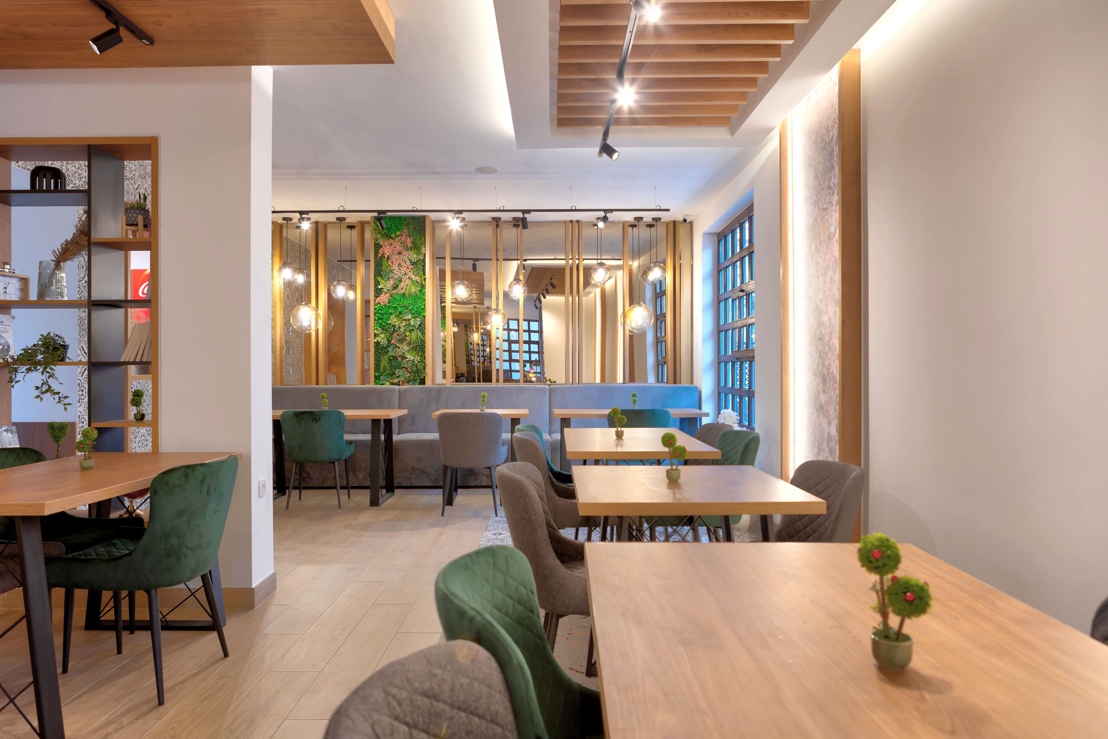 A modern dining area with wooden tables and green upholstered chairs, a green wall with a vertical garden, and a view of the city through large windows.